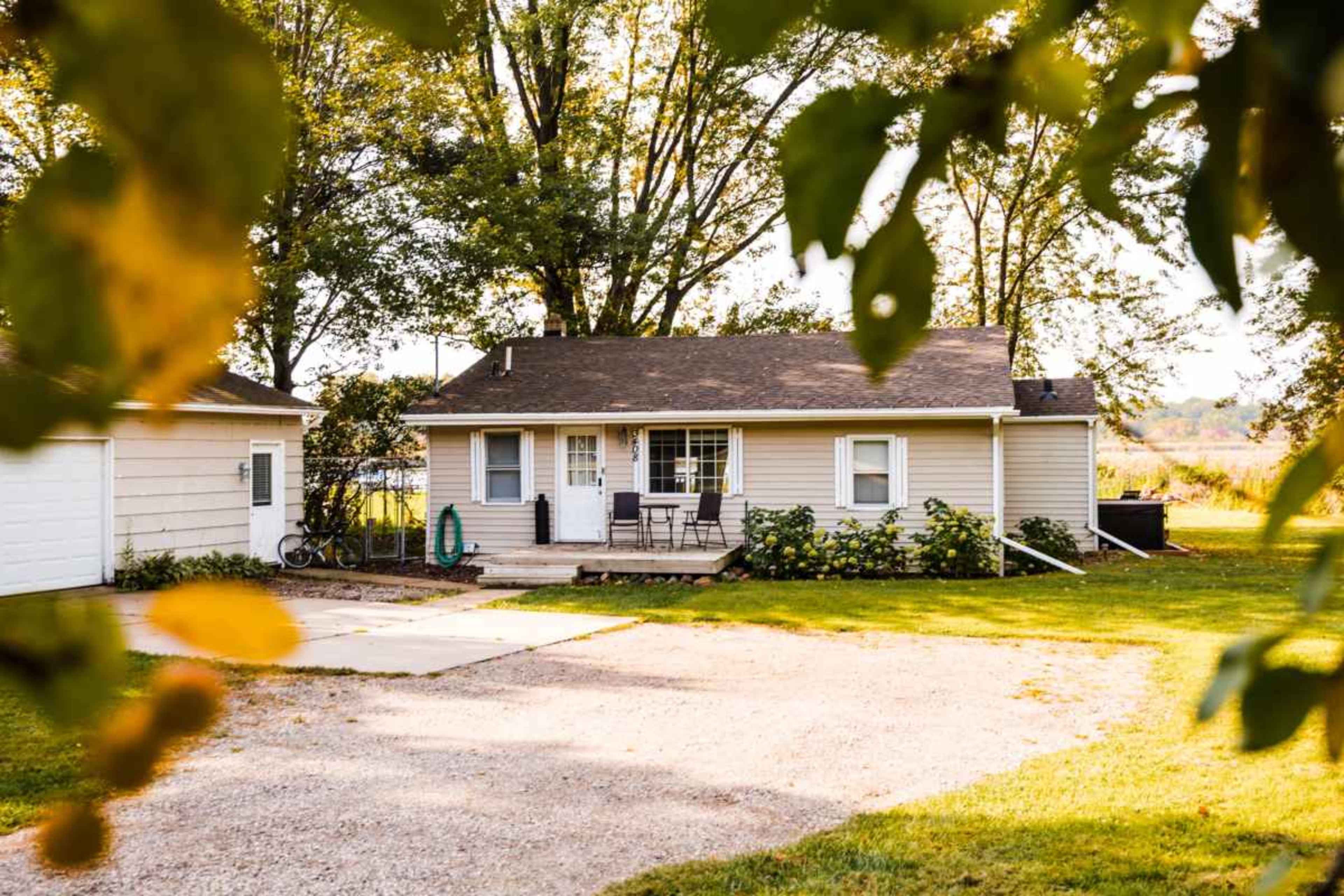A small, single-story house is set in a grassy area, surrounded by trees and a gravel driveway, with a porch and outdoor seating visible.