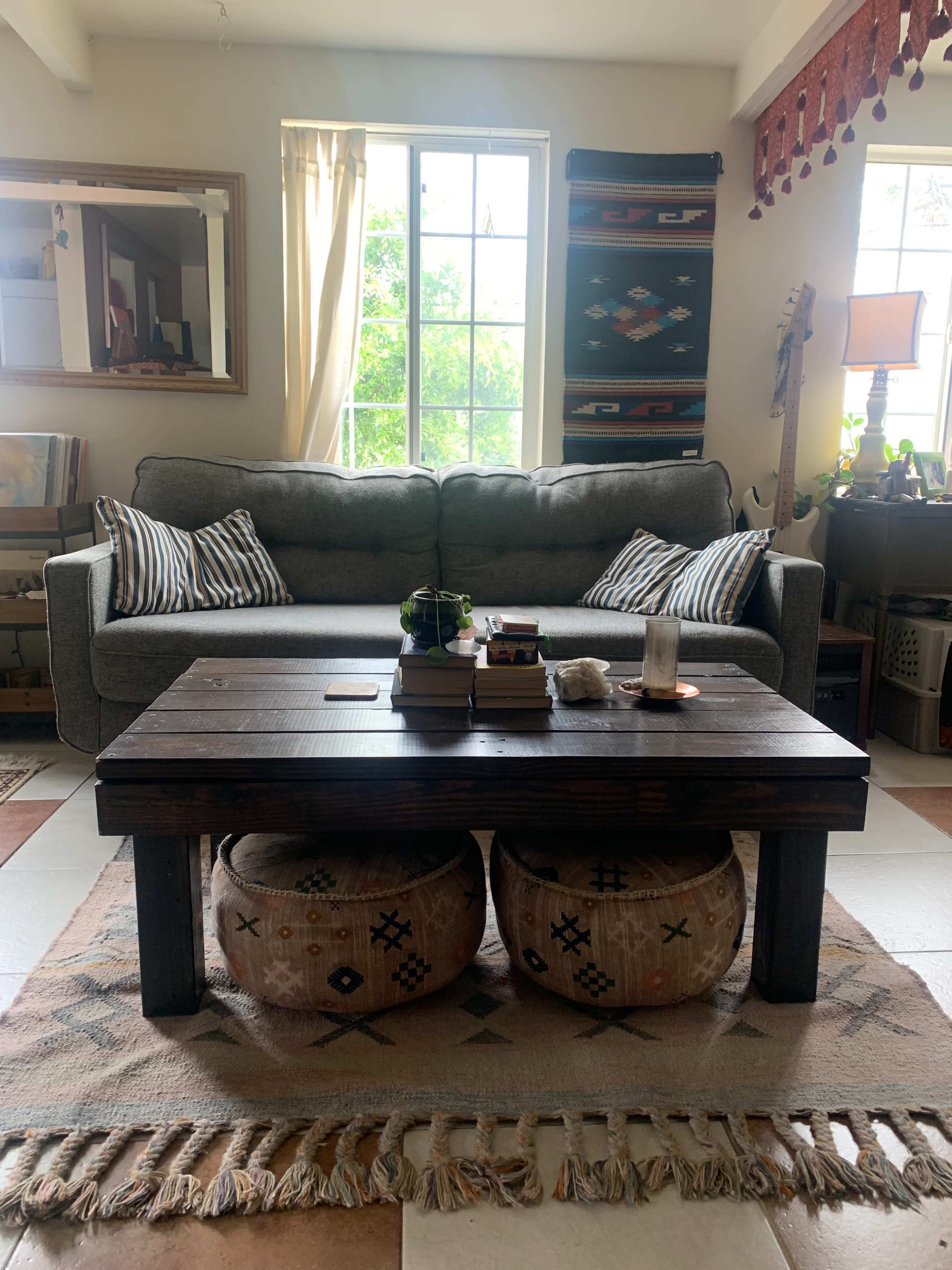 A living room with a gray sofa, a wooden coffee table, two patterned ottomans, and a window with greenery visible outside.