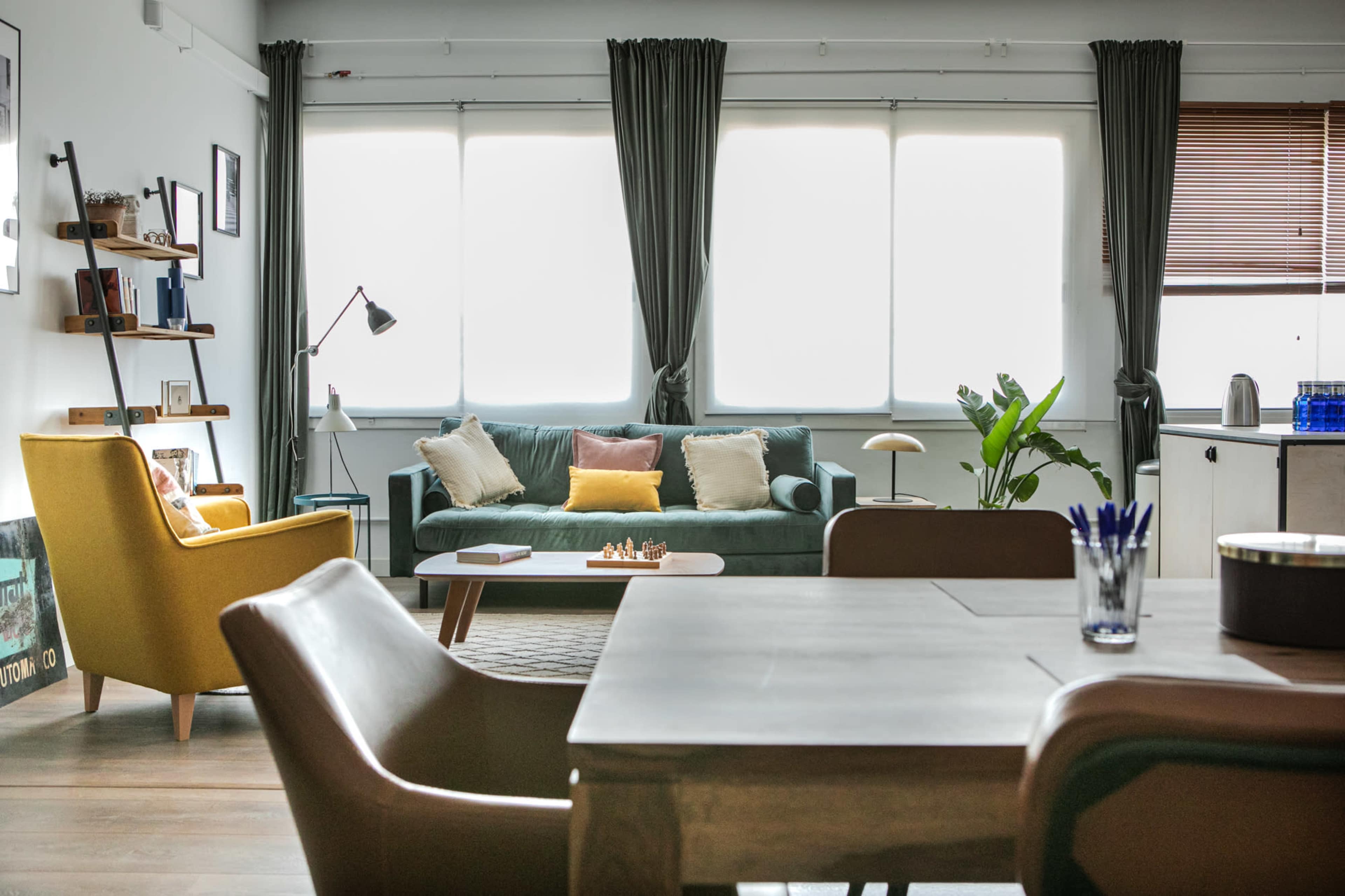 A modern living room with a green sofa, a yellow armchair, a dining table, and large windows covered by curtains.
