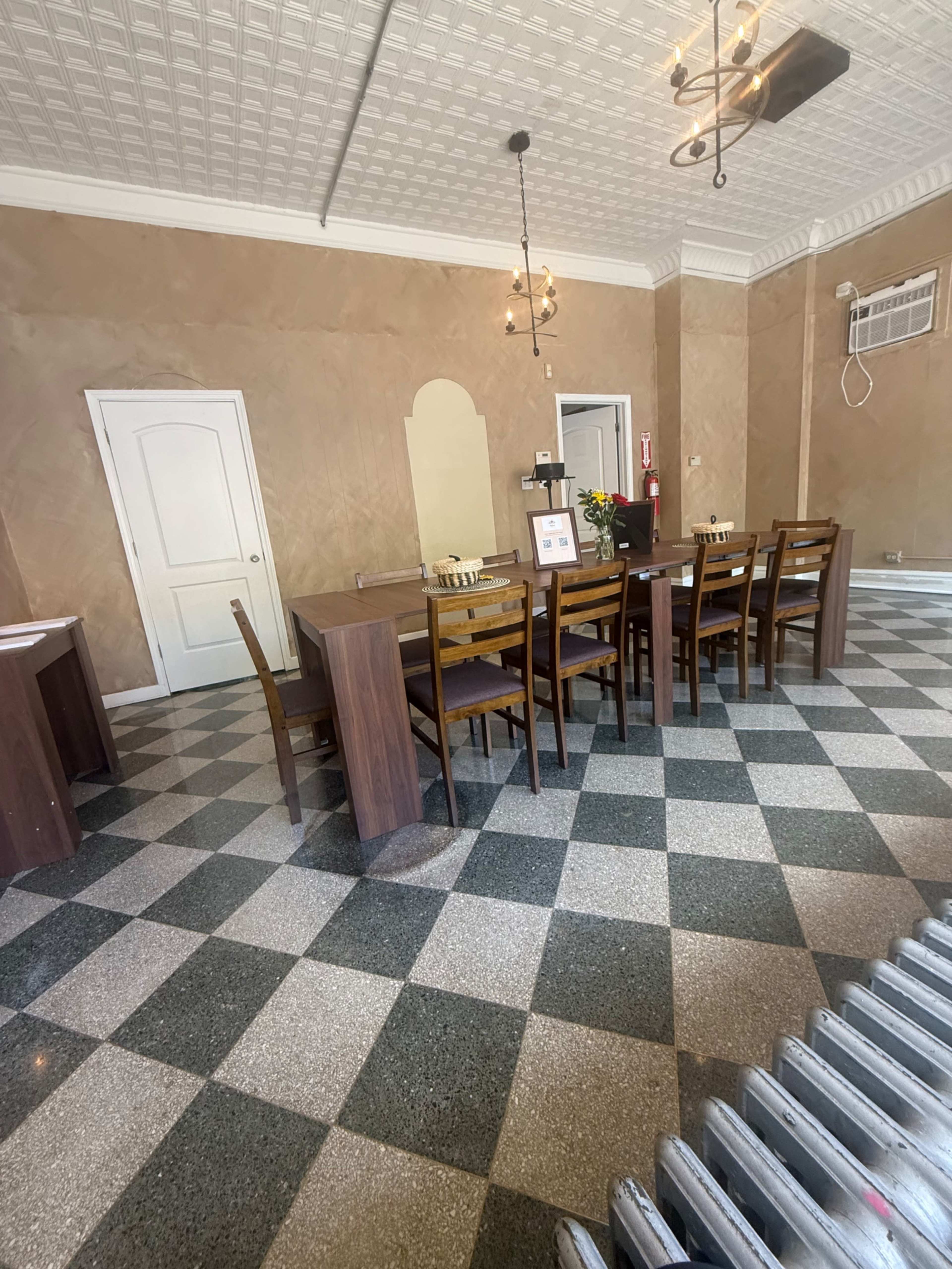 The image shows a dining area with a long wooden table surrounded by chairs, set against a backdrop of textured walls and a checkerboard floor.