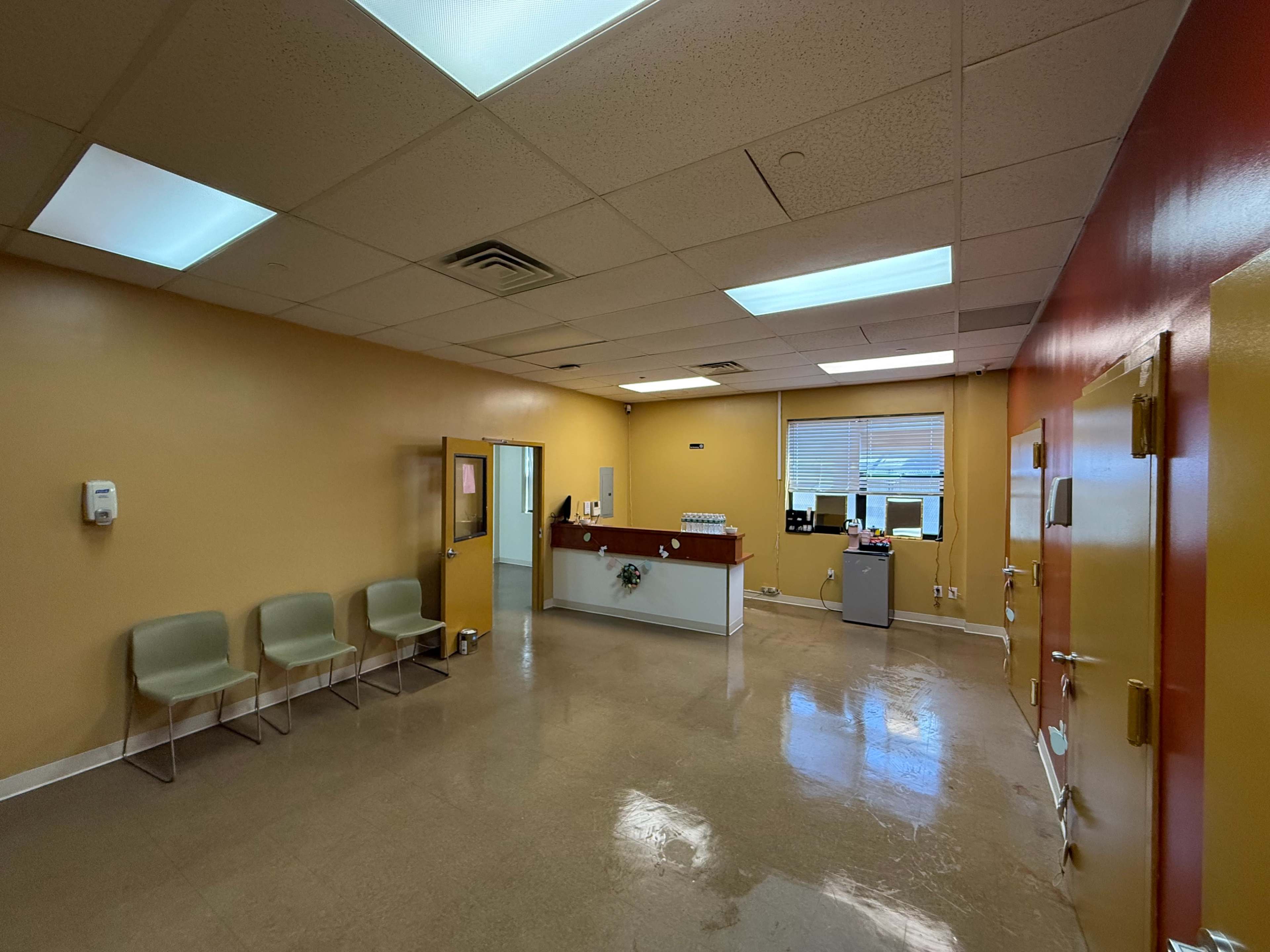 The image shows a brightly lit waiting area of an office, featuring a reception counter, a few chairs, and a workstation in the background.