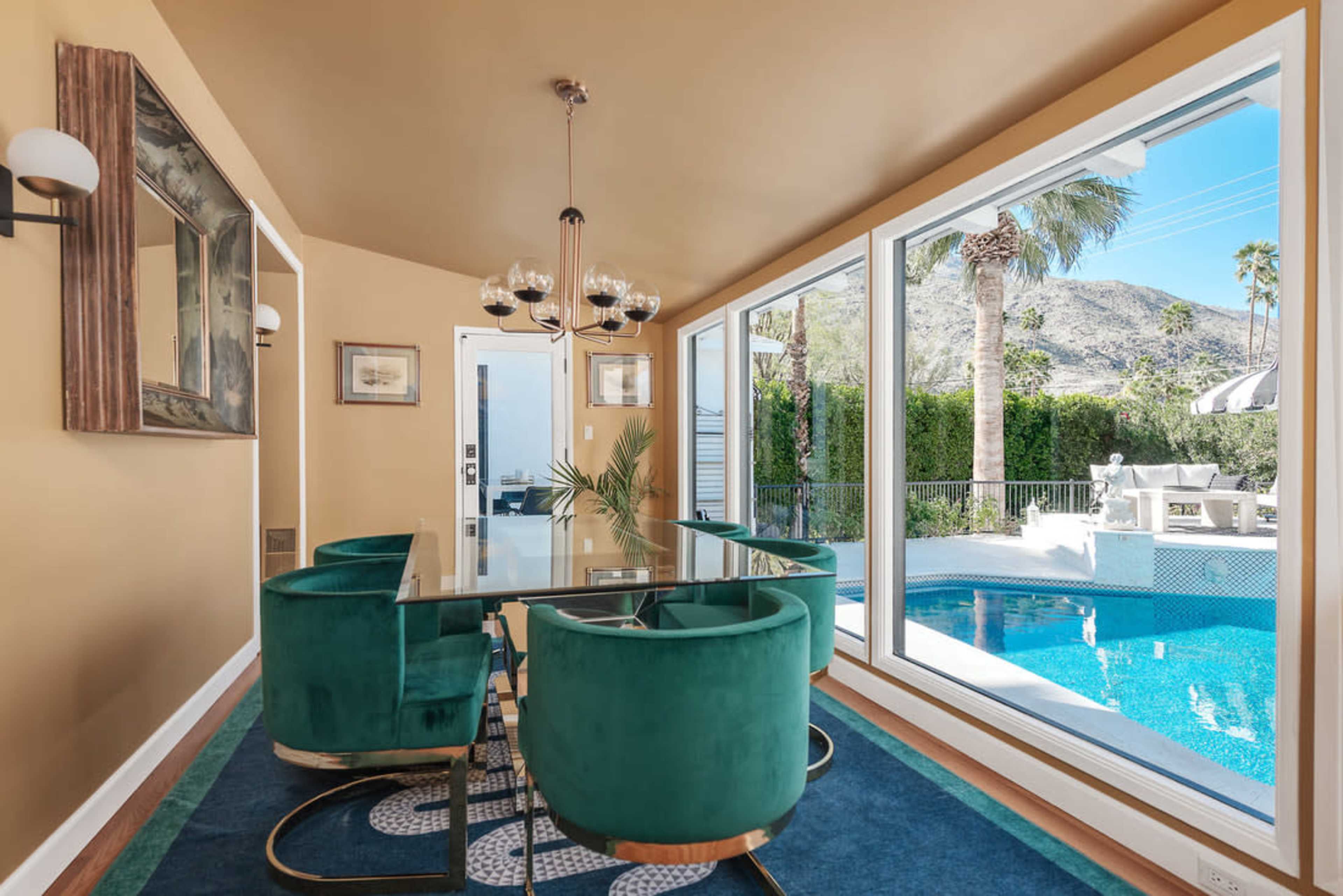 A modern dining area features a glass table surrounded by four green velvet chairs, with large windows offering views of a pool and palm trees outside.