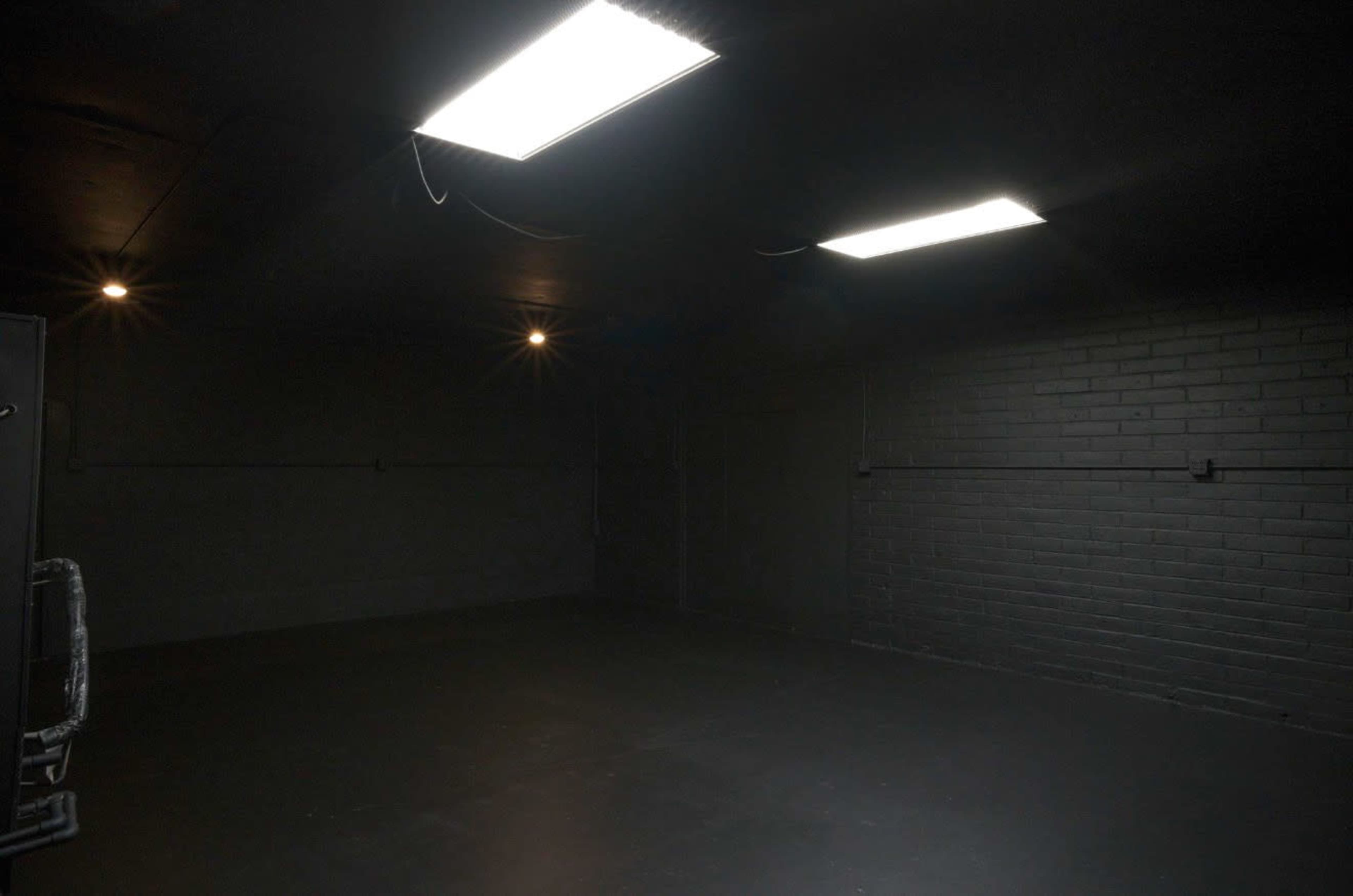 The image shows a dimly lit, empty room with black walls, a concrete floor, and overhead fluorescent lights.