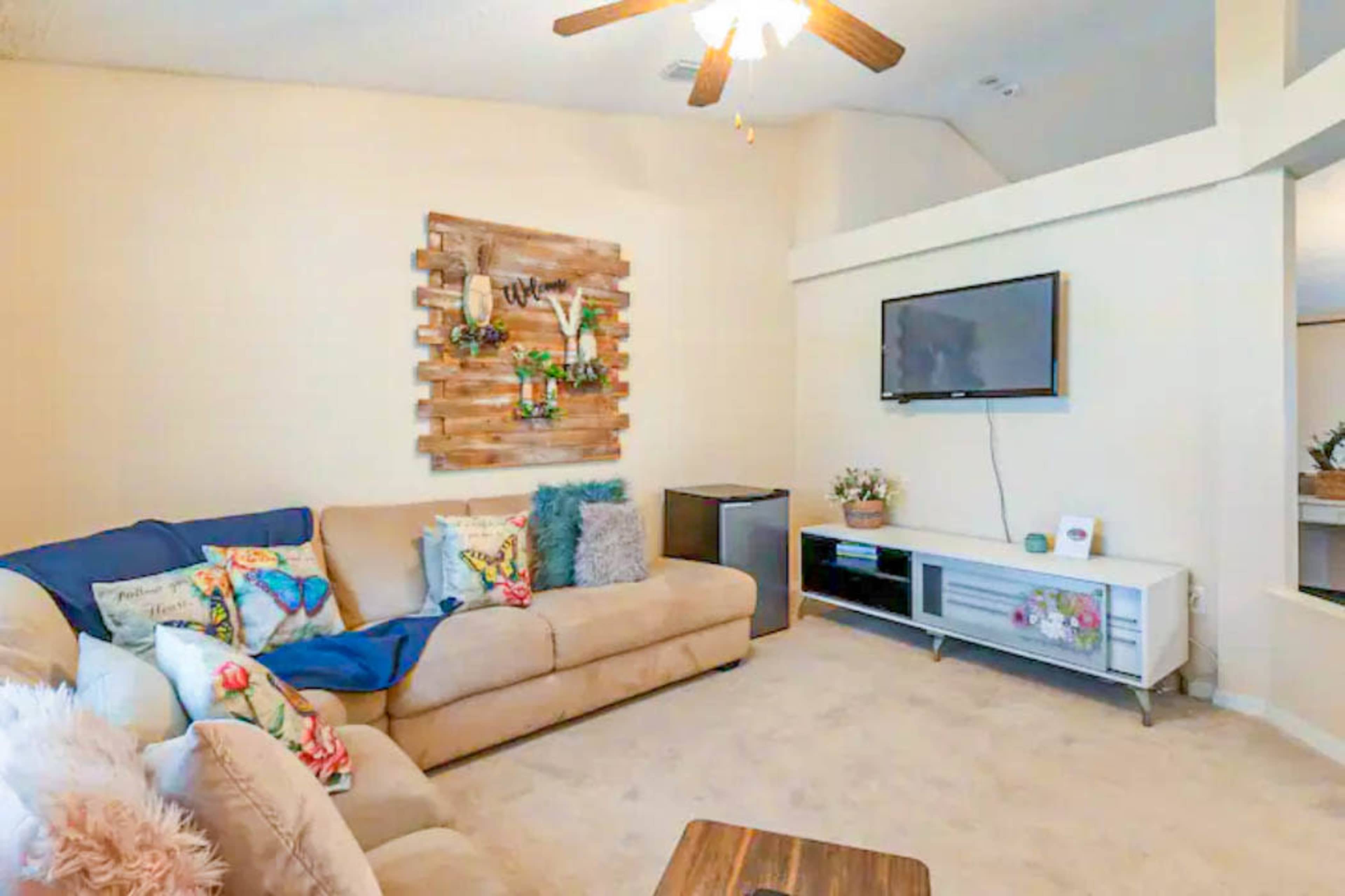A cozy living room features a beige couch with colorful pillows, a wooden wall decoration with plants, a mounted television, and a light-colored carpeted floor.