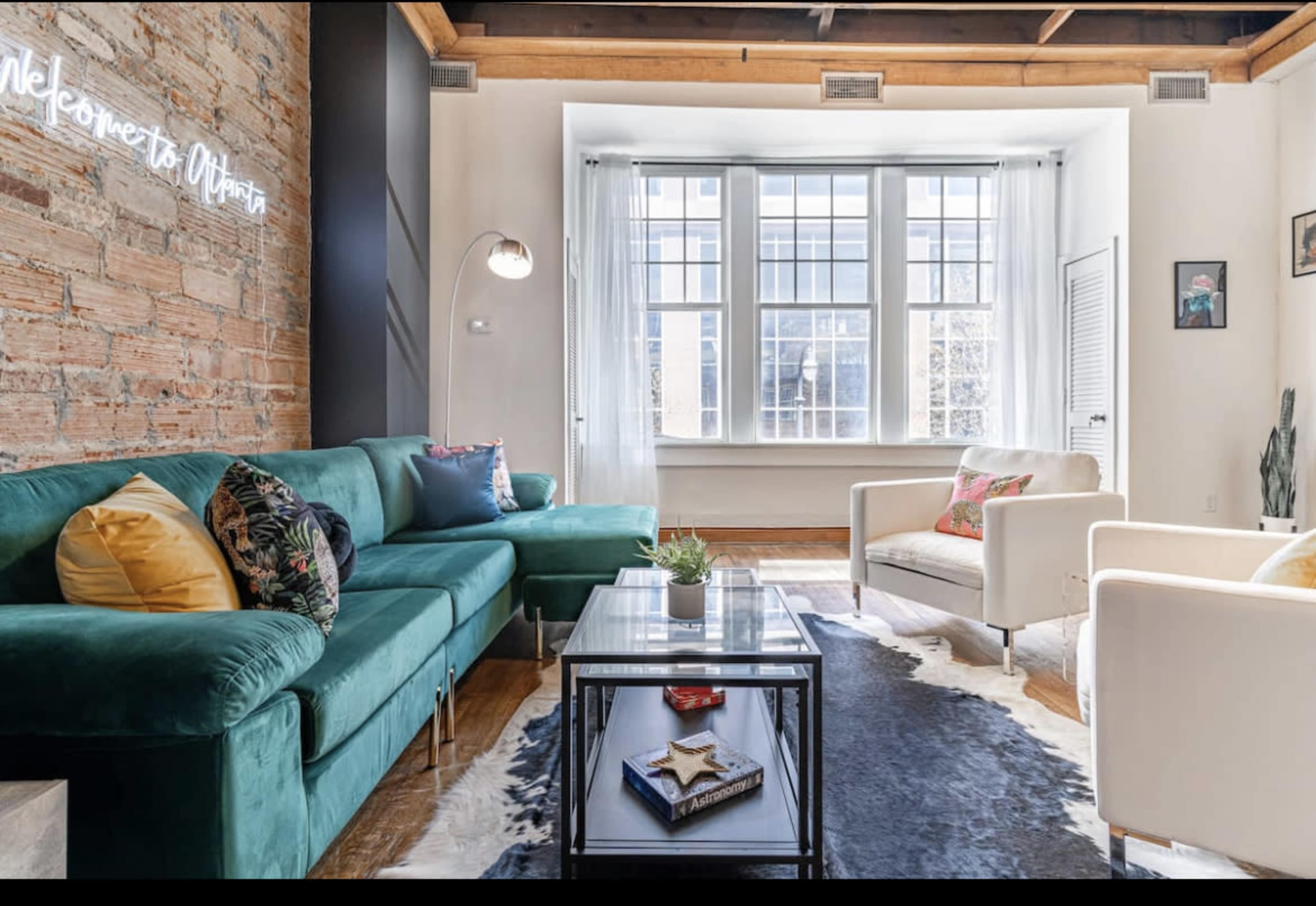 The living room features a green velvet sofa, a white armchair, a glass coffee table, and large windows allowing natural light to fill the space.