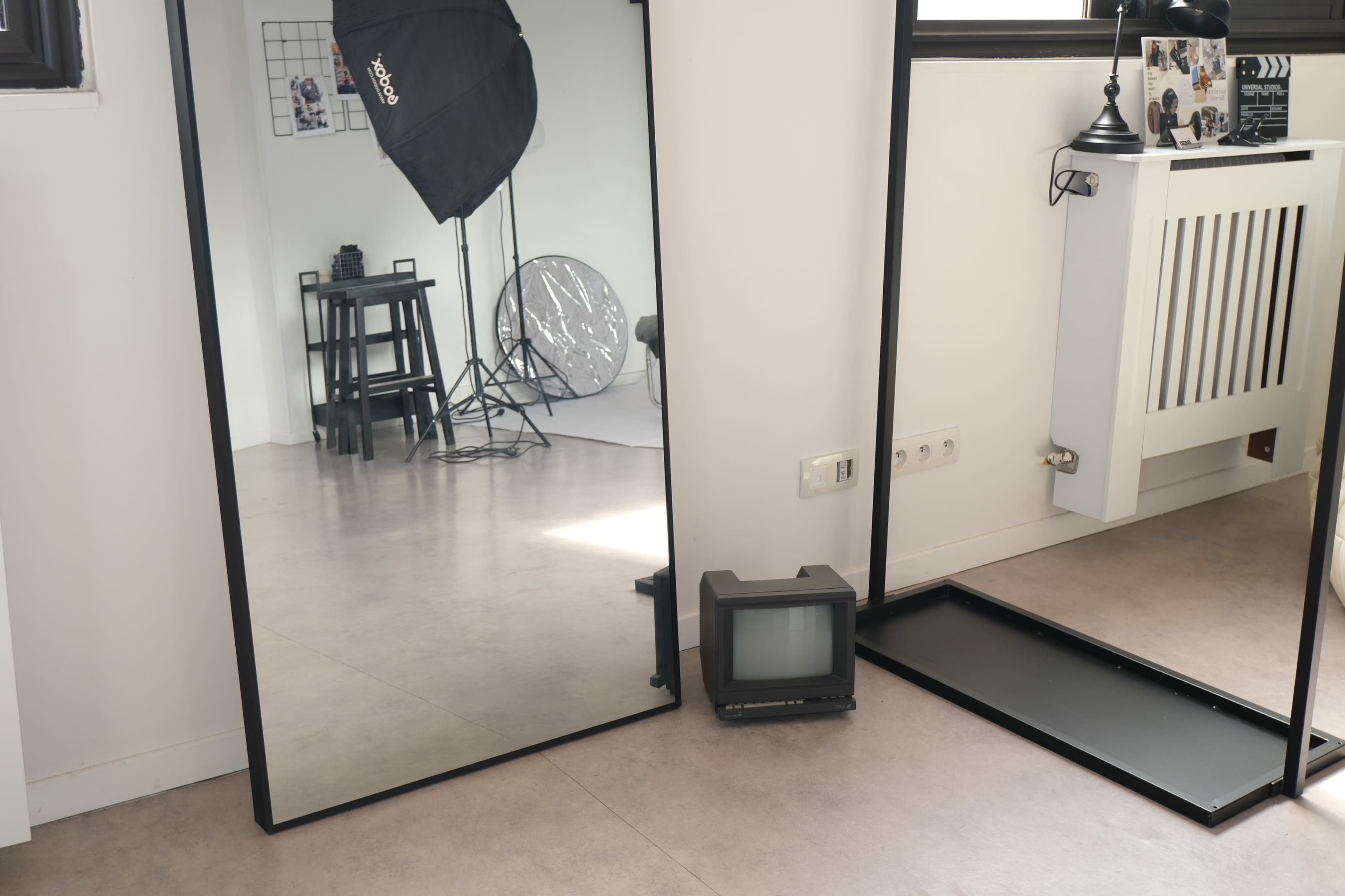 A small vintage television is positioned beside a tall mirror reflecting a studio space with a stool and softbox in the background.