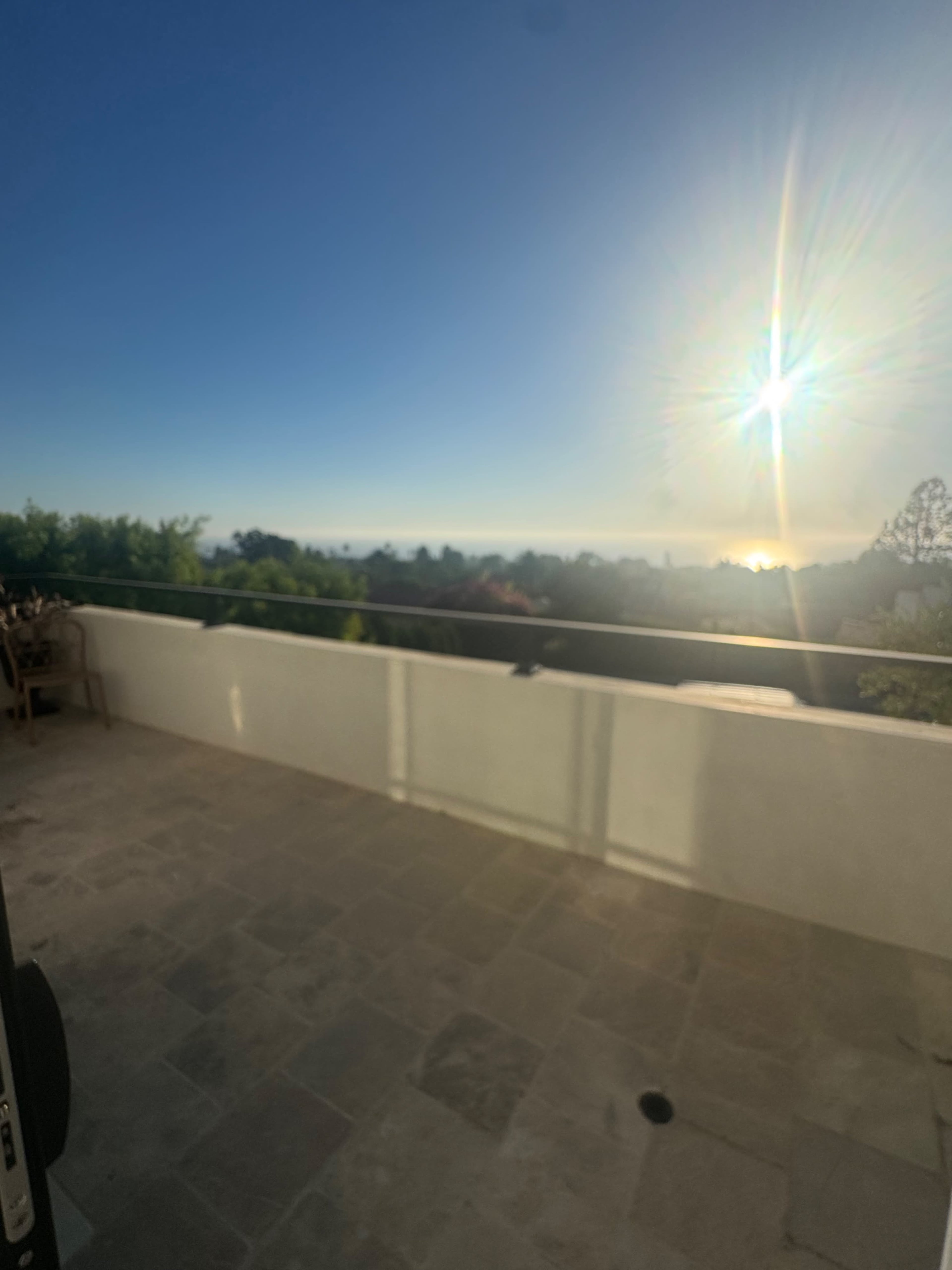 A bright sun sets over a distant horizon, viewed from a stone terrace surrounded by greenery.