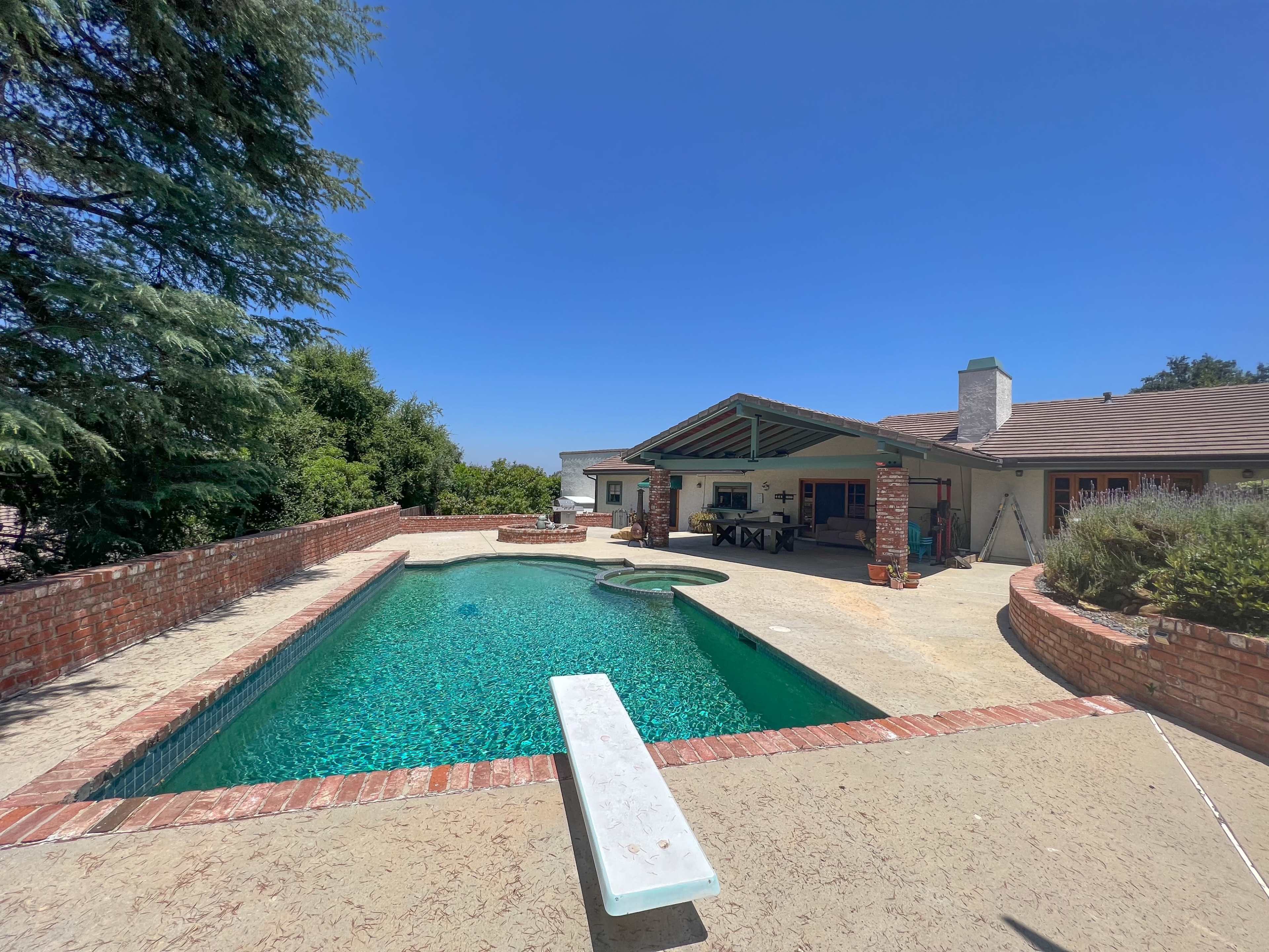 A backyard patio with a swimming pool, surrounded by brick walls and green foliage under a clear blue sky.