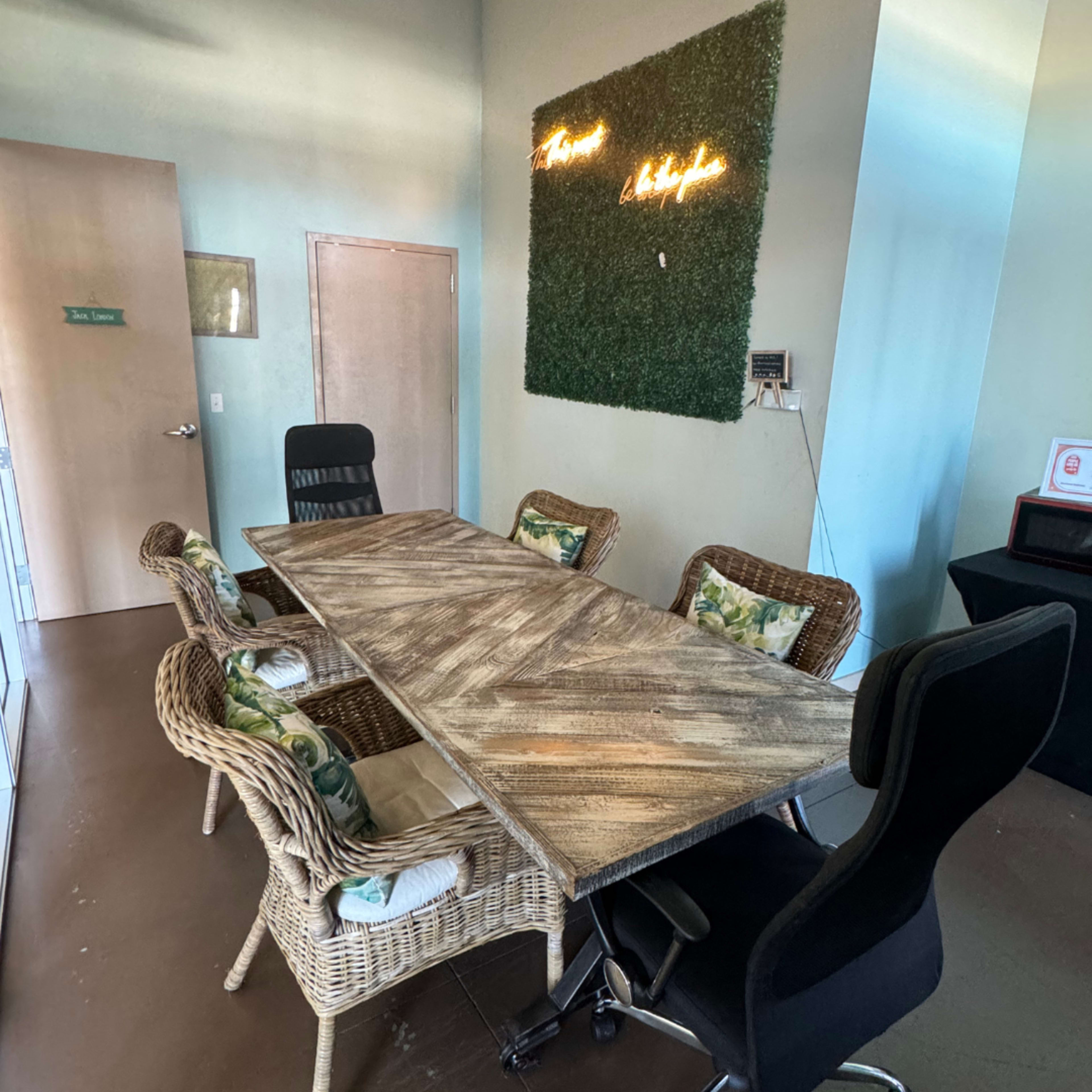The image shows a modern conference room with a wooden table surrounded by wicker and black chairs, featuring a green wall decor with neon text.