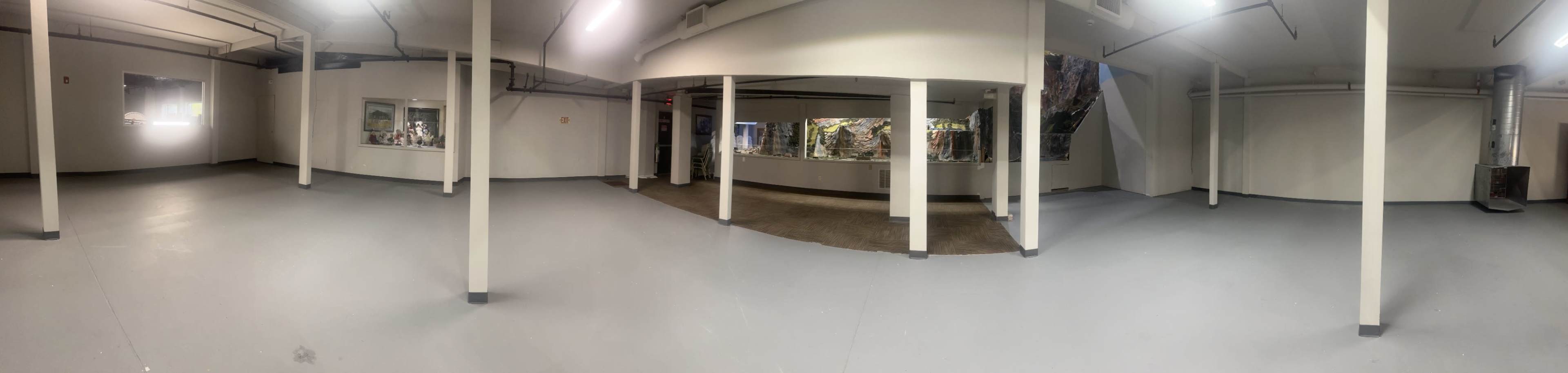 The image shows a spacious, vacant interior room with grey flooring and white walls, featuring several columns and large windows along one side.