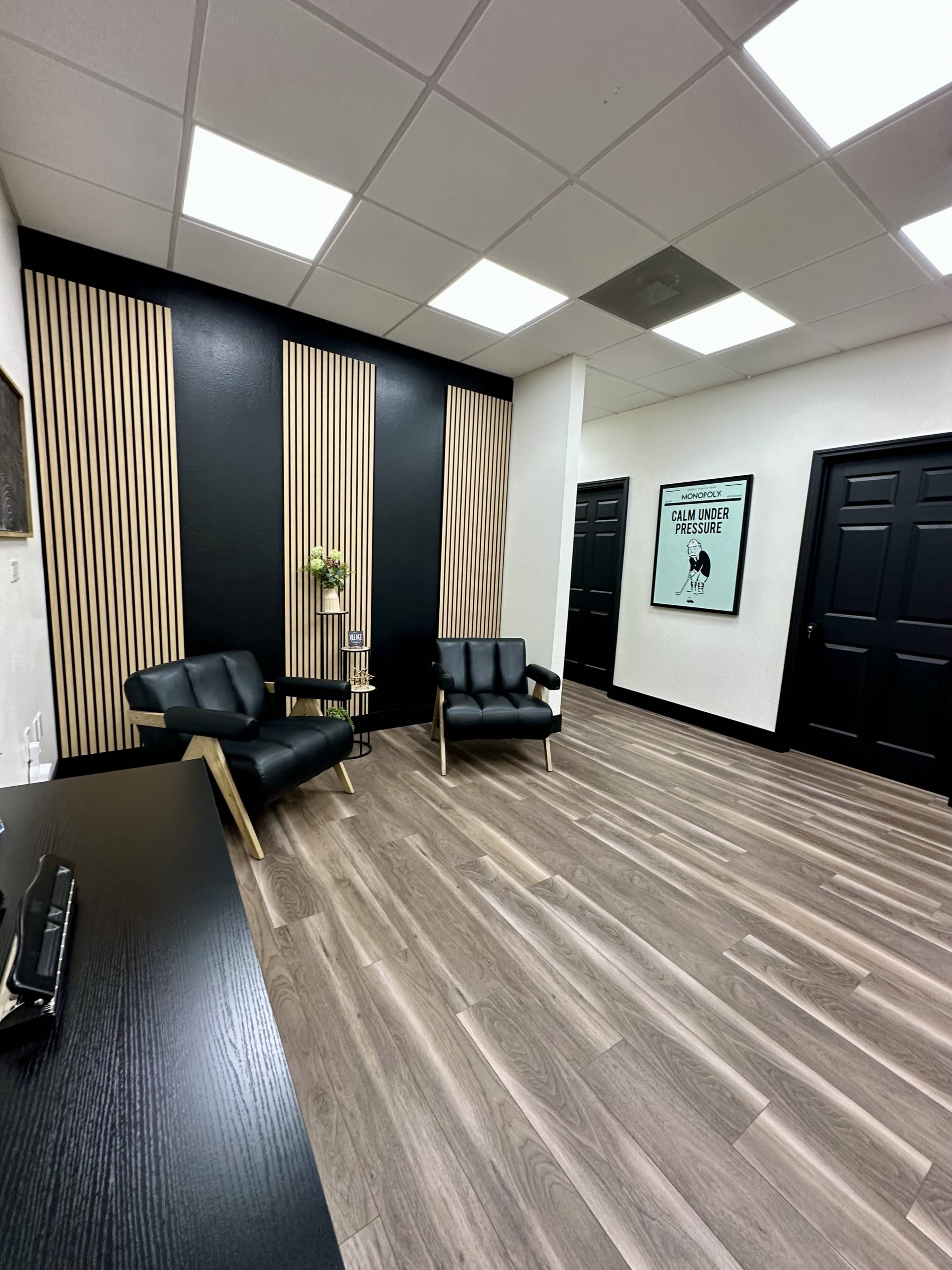The image shows a modern office waiting area featuring two black leather chairs, a small round table, and wood-paneled accent walls.