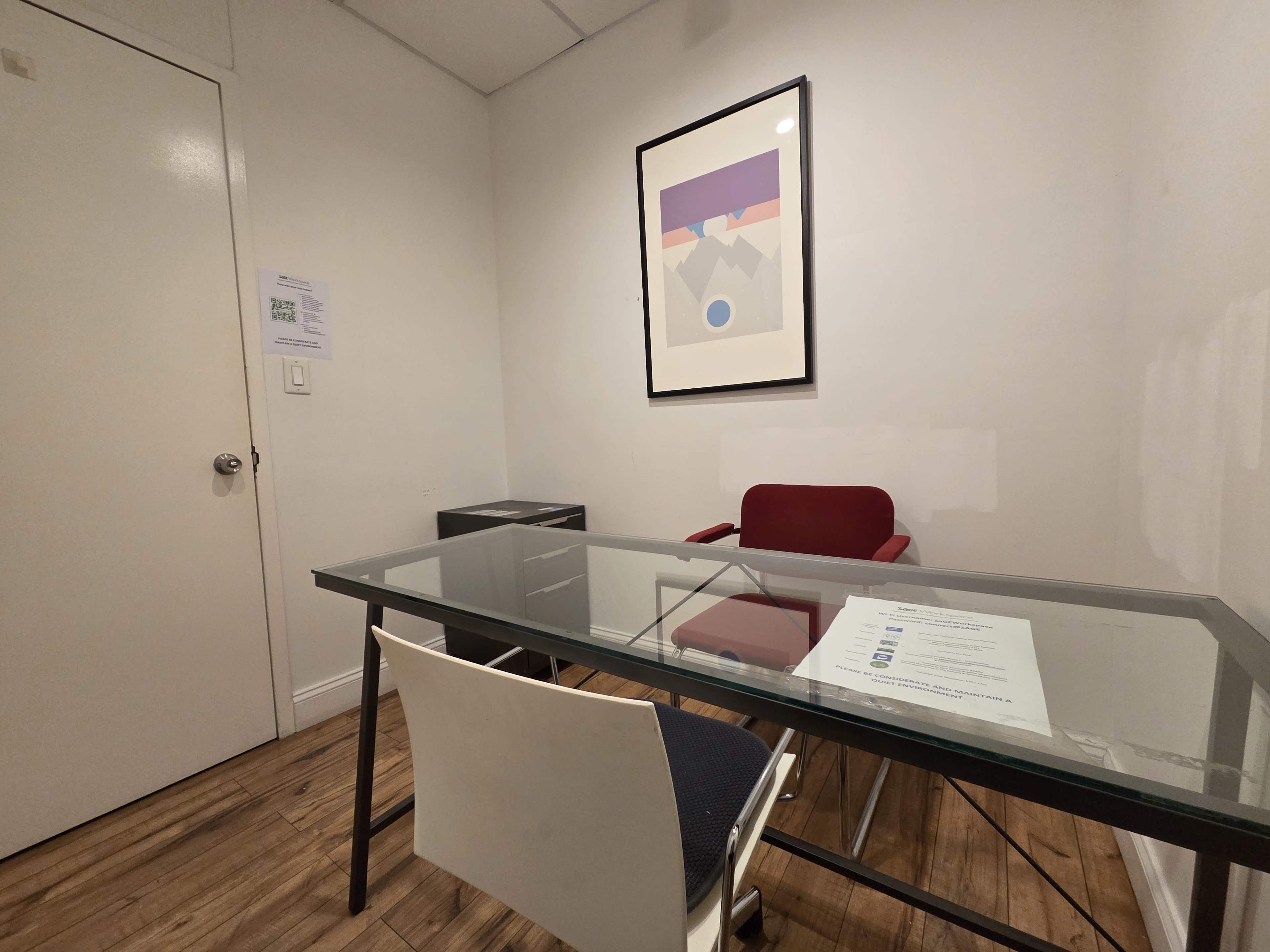 The image shows a small office with a glass table, a red chair, and a framed artwork on the wall.