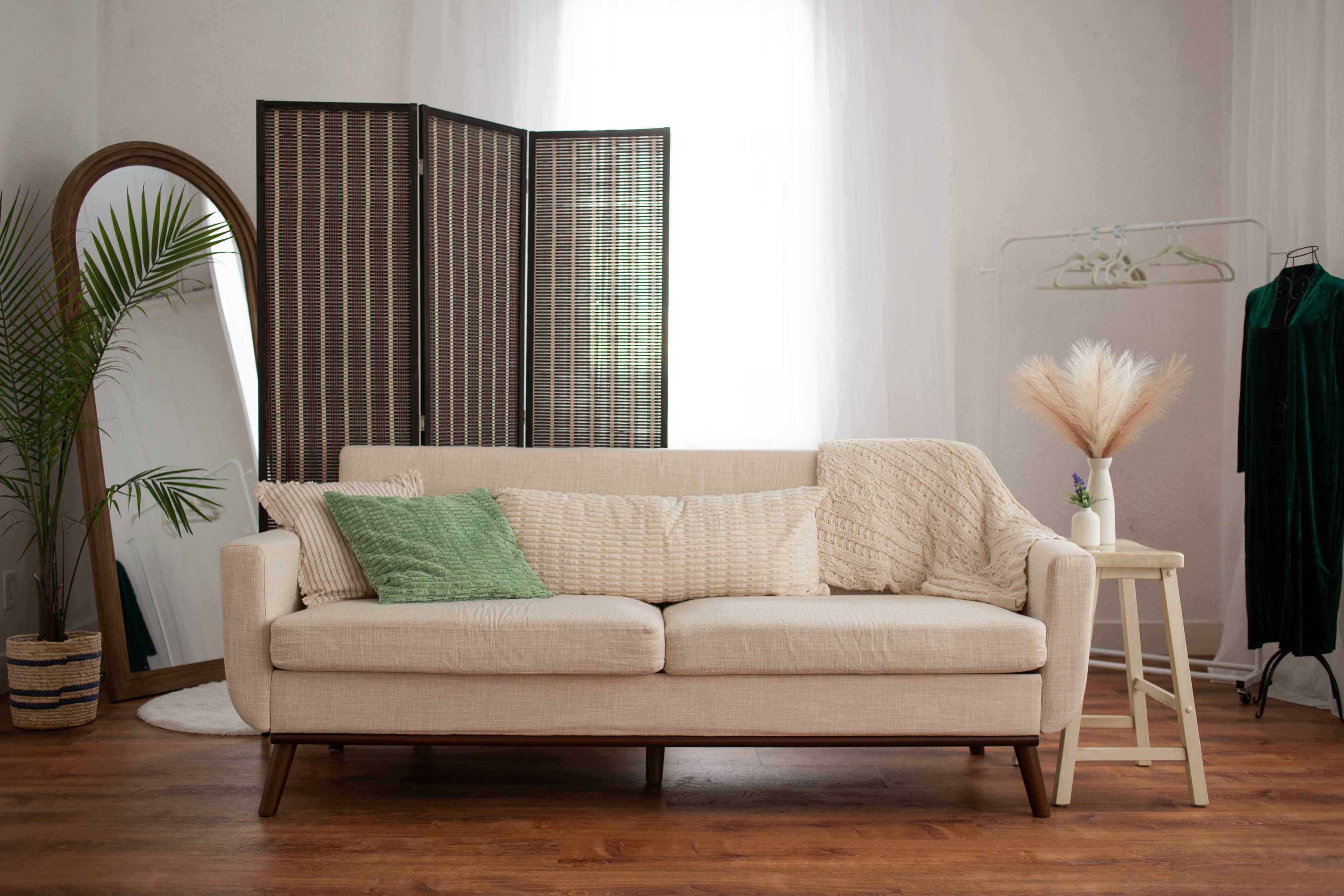 The image features a light-colored sofa adorned with various pillows, positioned beside a tall mirror and a folding screen in a well-lit room with wooden flooring.