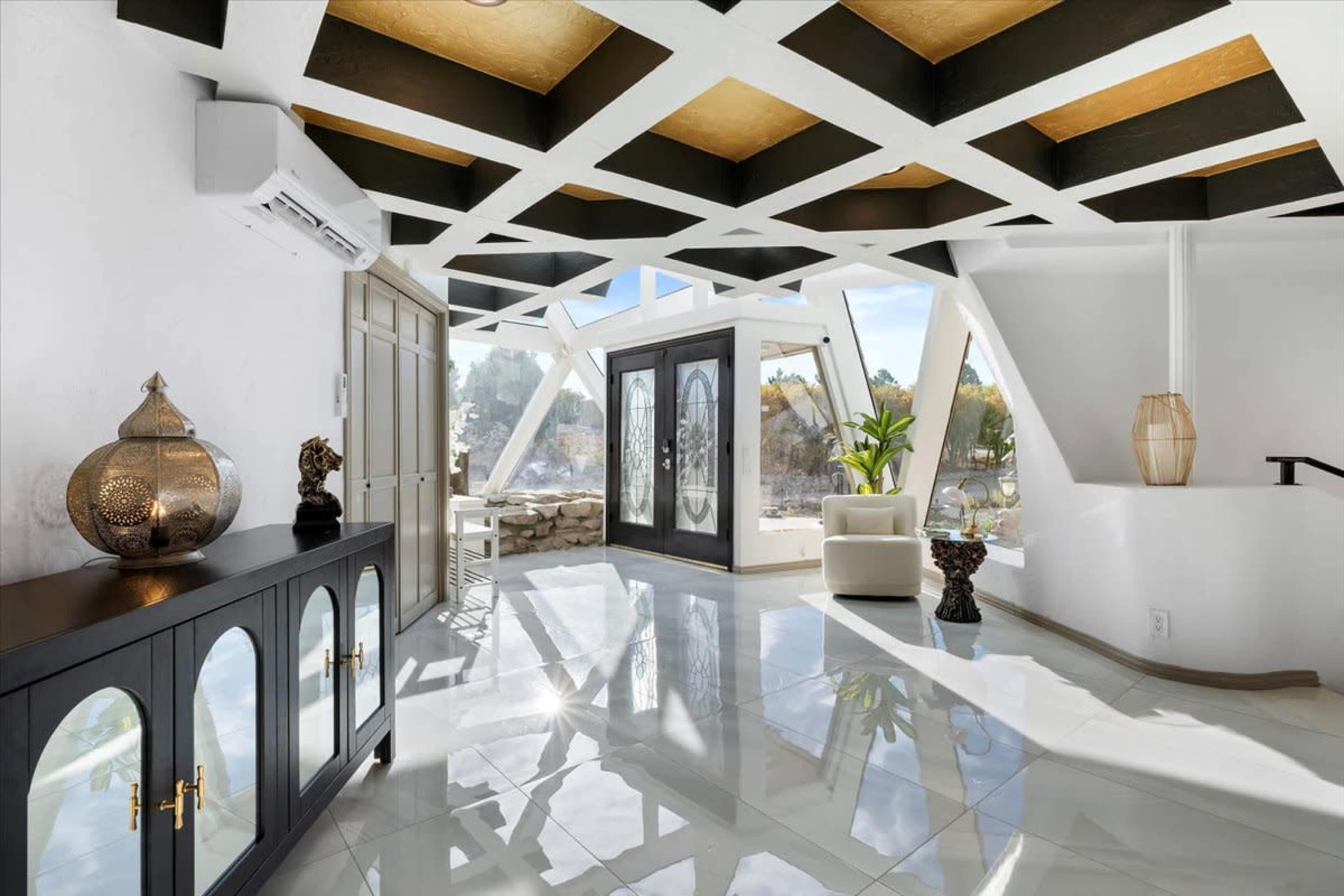 The image depicts a modern interior space with a geometric ceiling design, large windows allowing natural light, and polished tile flooring.