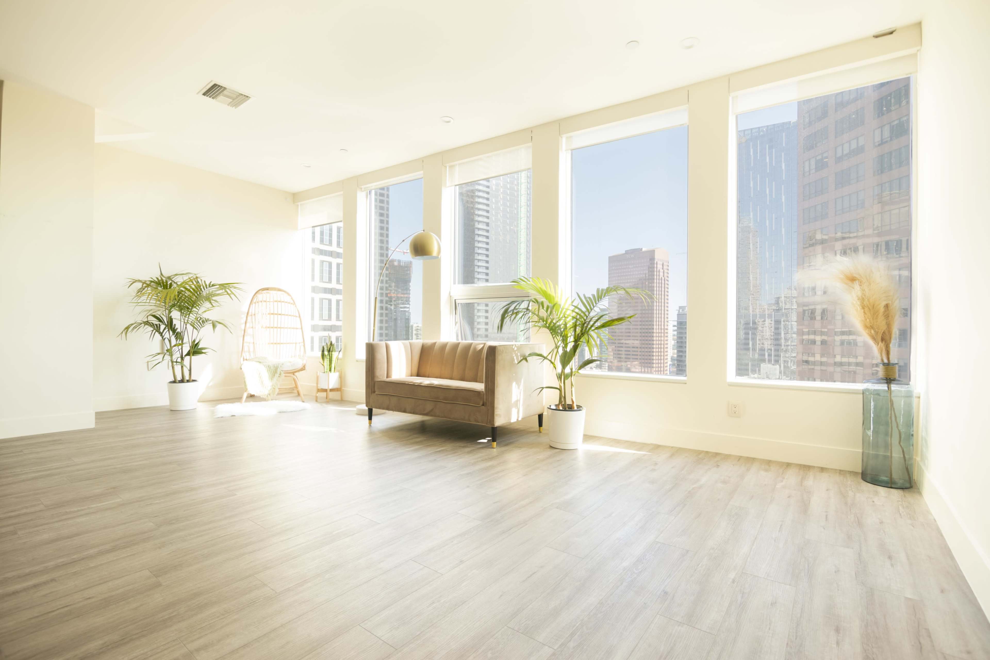 A spacious, well-lit room features a sofa and large windows with city views, surrounded by potted plants.