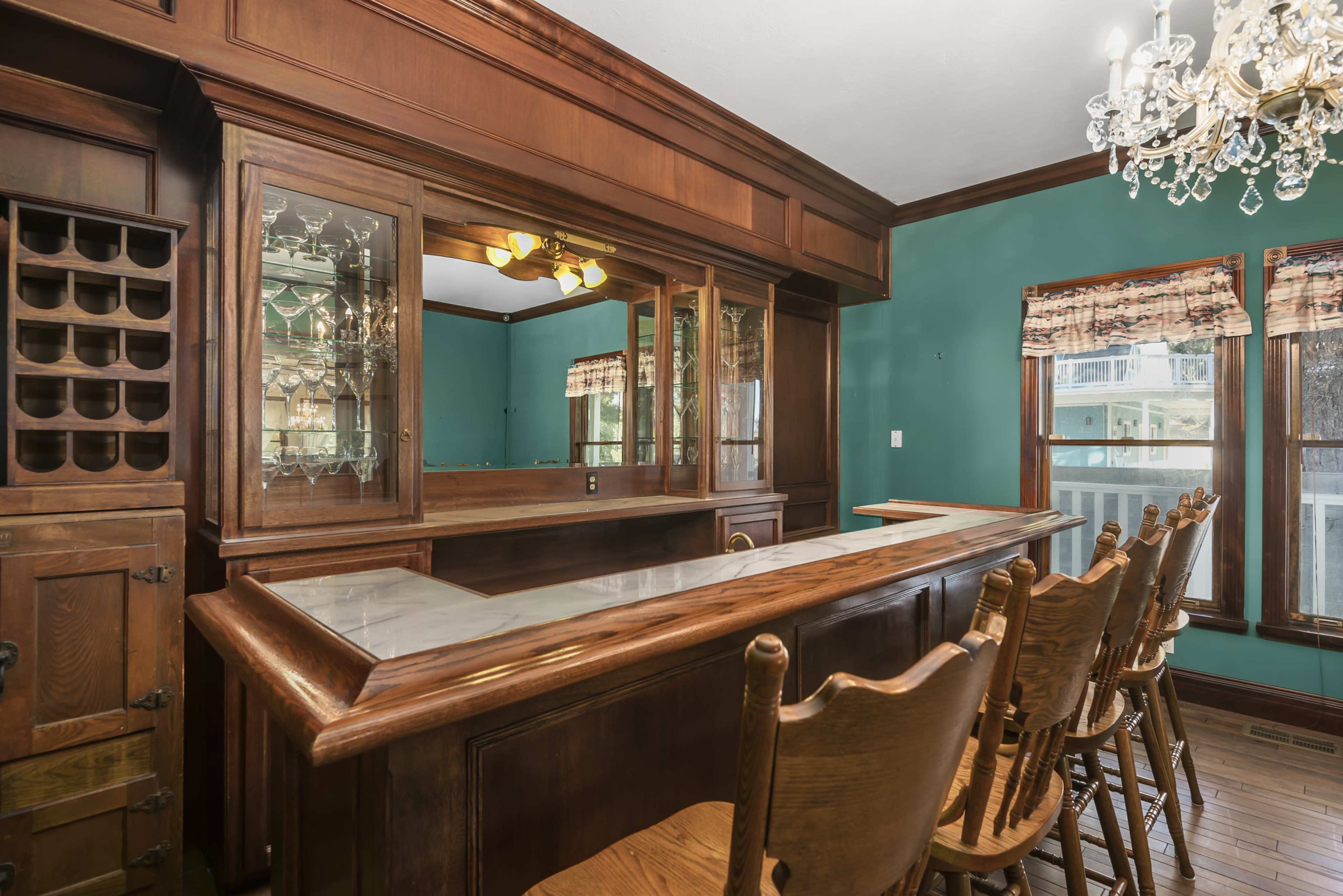 A wooden home bar with a polished countertop, backlit glass shelves, and several bar stools arranged in front of it.