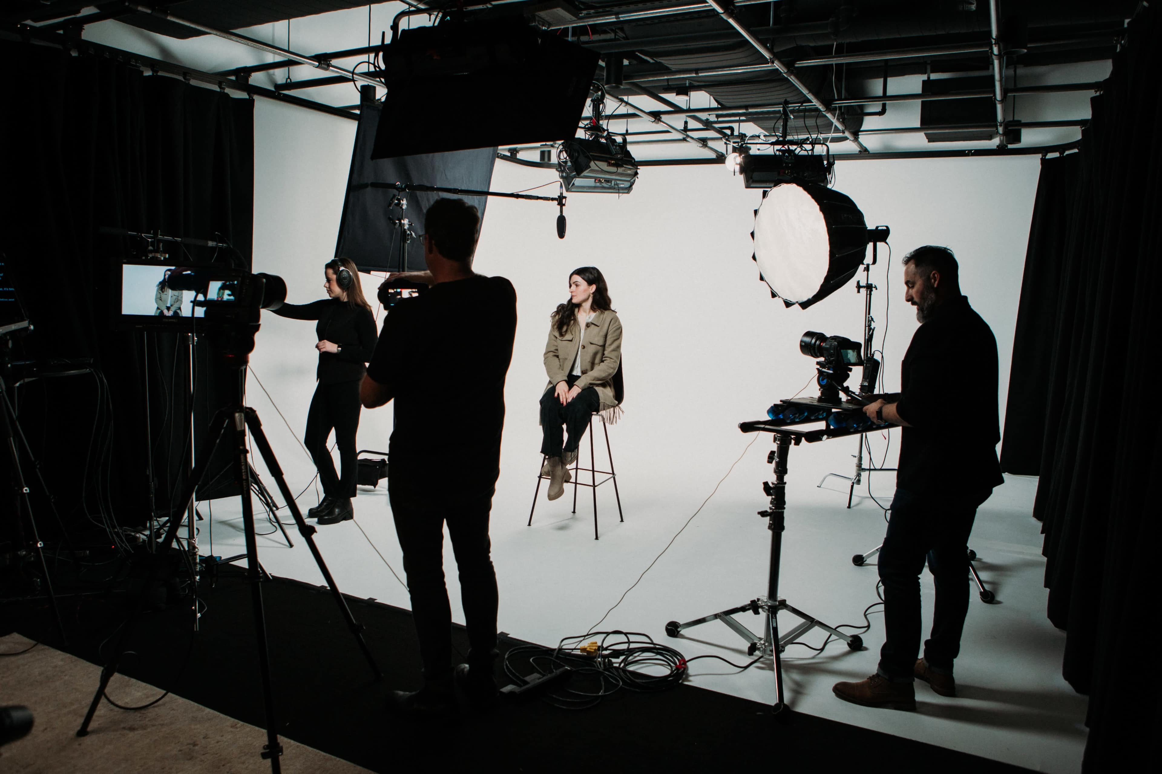 A film crew sets up equipment around a woman seated on a stool in a bright studio with a white backdrop.