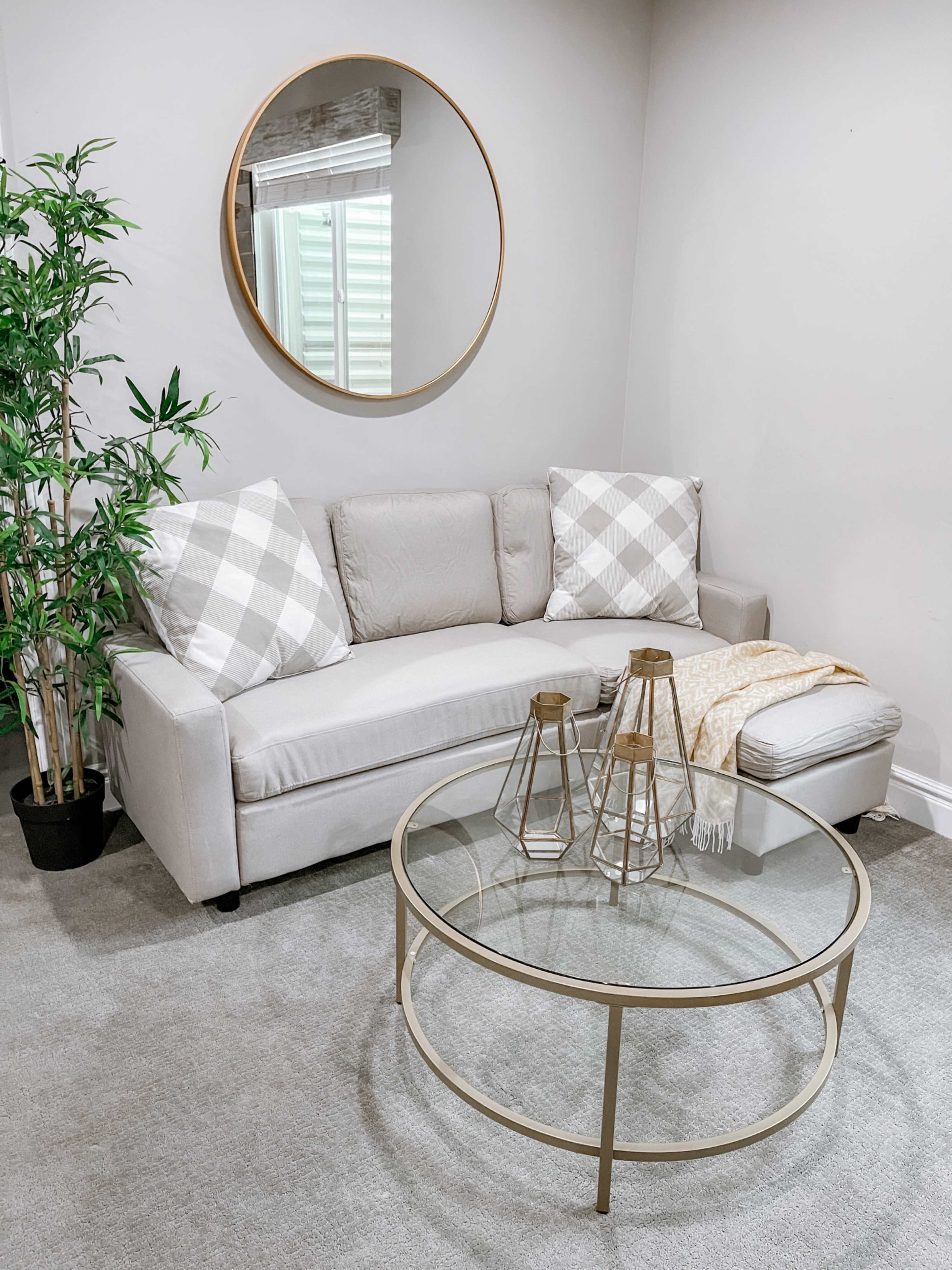 The image shows a light-colored sofa with patterned cushions, a round mirror on the wall, a glass coffee table with decorative items, and a potted plant in a corner.