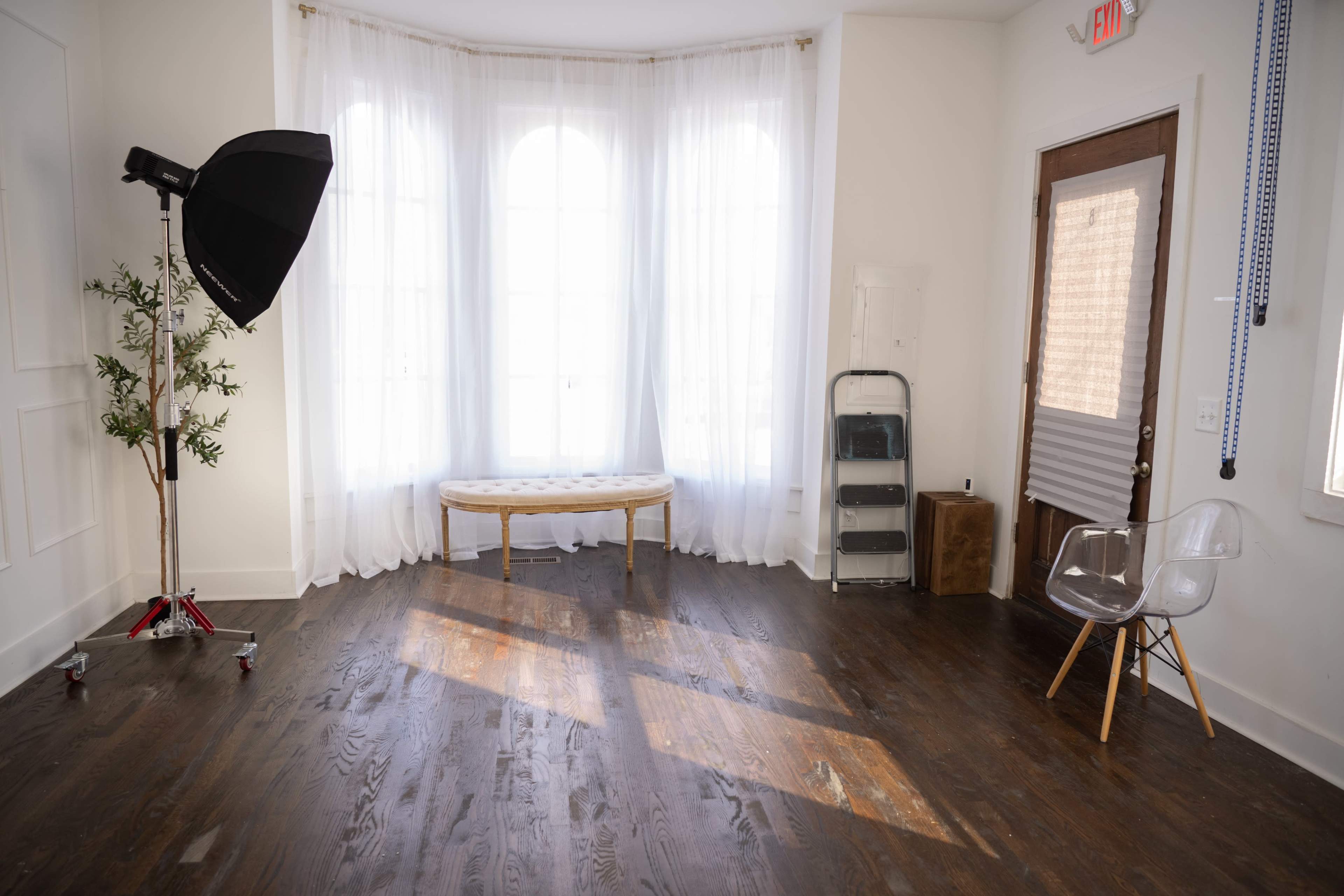The image shows a well-lit room with large windows, a bench in the center, and a photography umbrella on a stand in one corner.