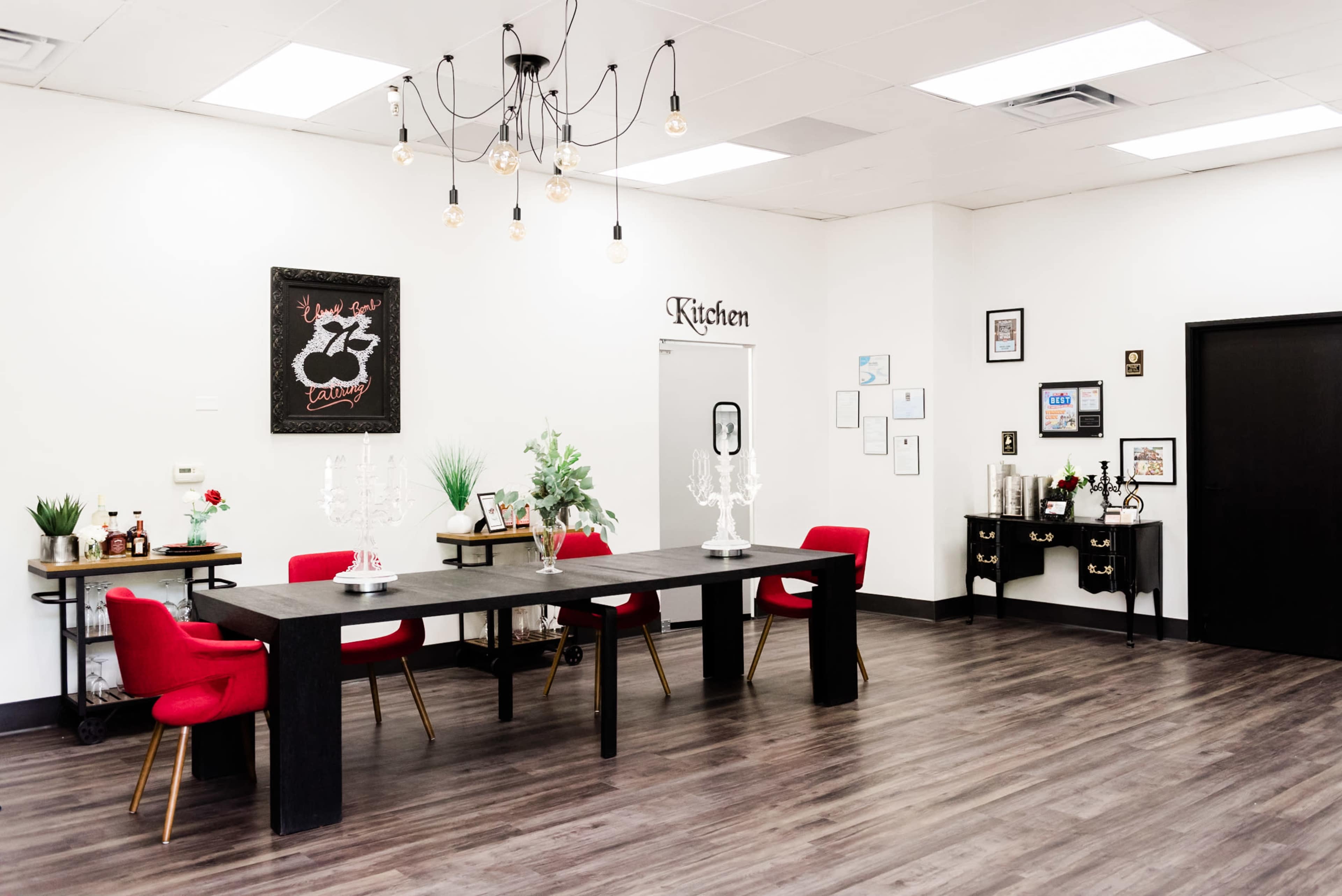 A modern, well-lit kitchen space featuring a long black table surrounded by red chairs, greenery, and decorative accents on the walls.