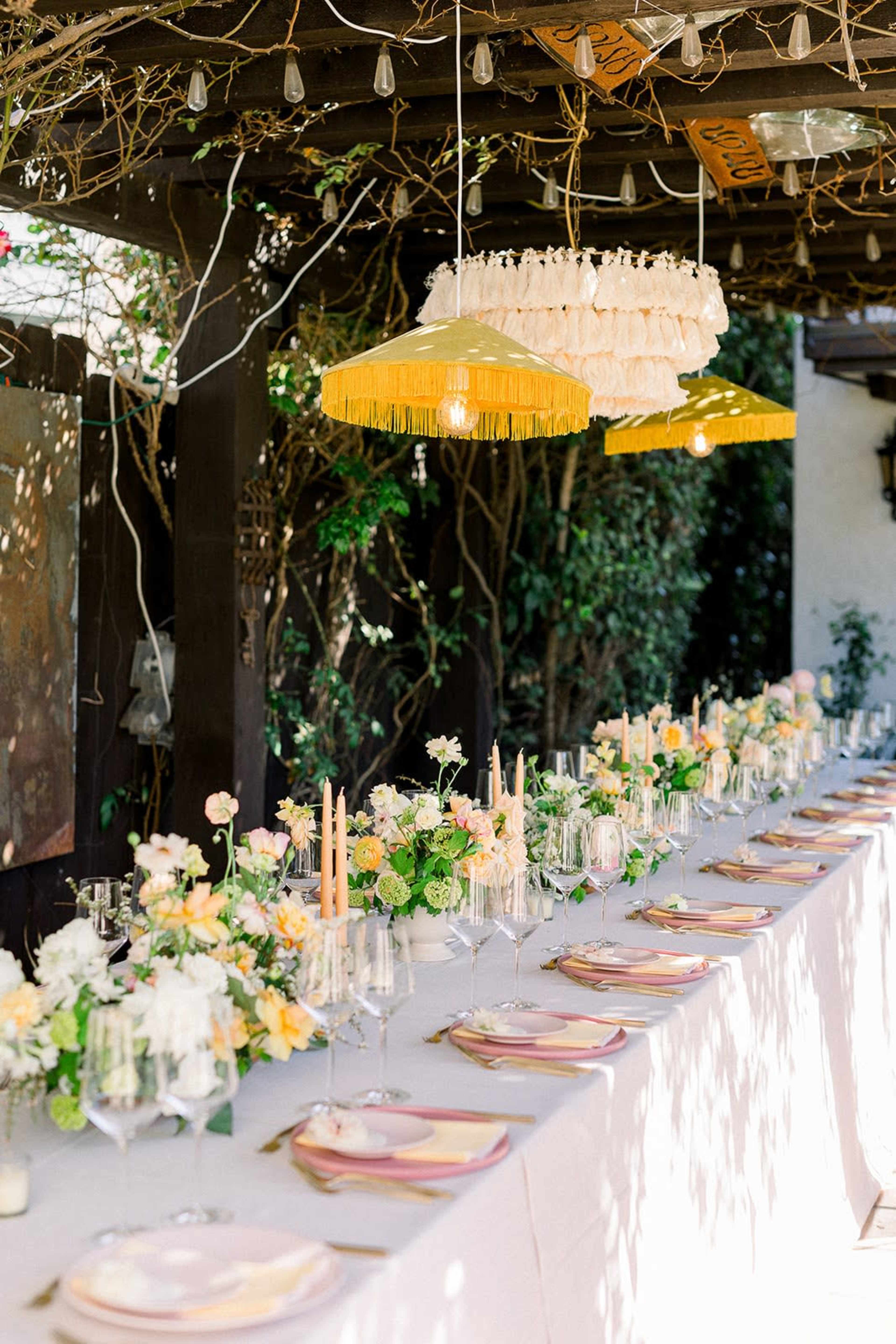 A long dining table elegantly set with floral centerpieces and candles is arranged under a pergola adorned with hanging light fixtures.