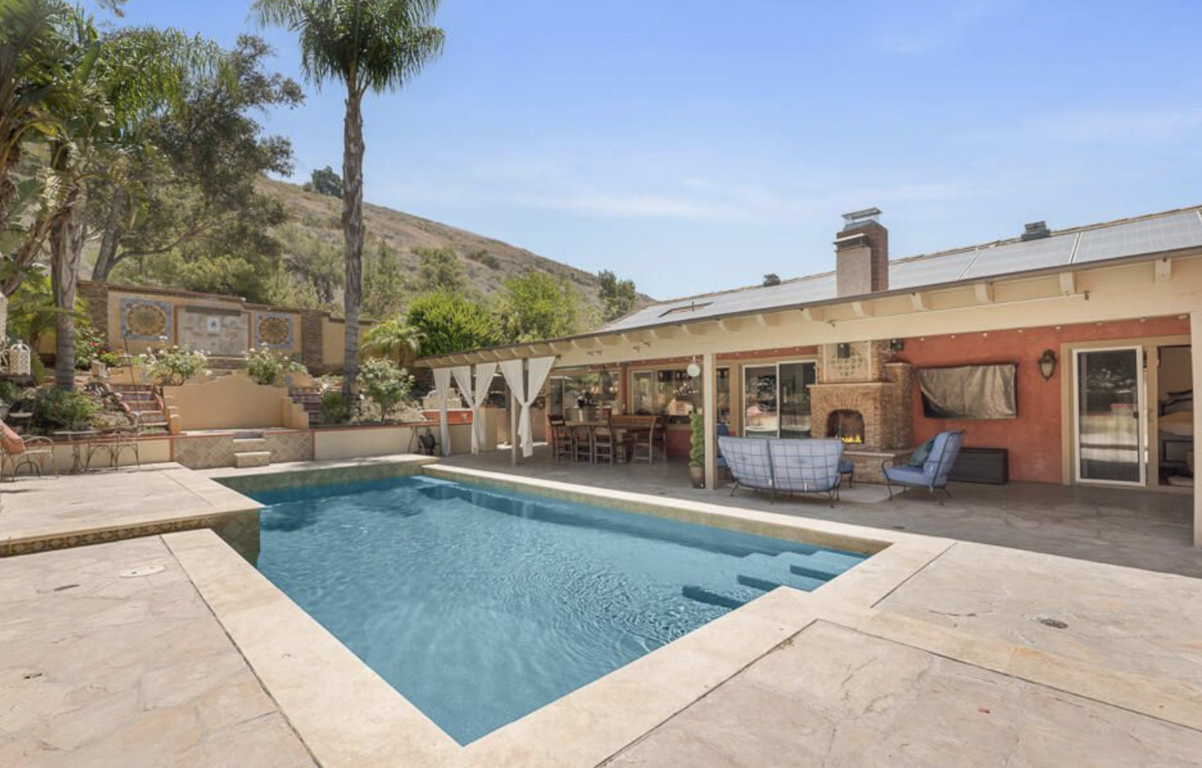 A pool area with a patio and outdoor seating adjacent to a house surrounded by trees and hills.