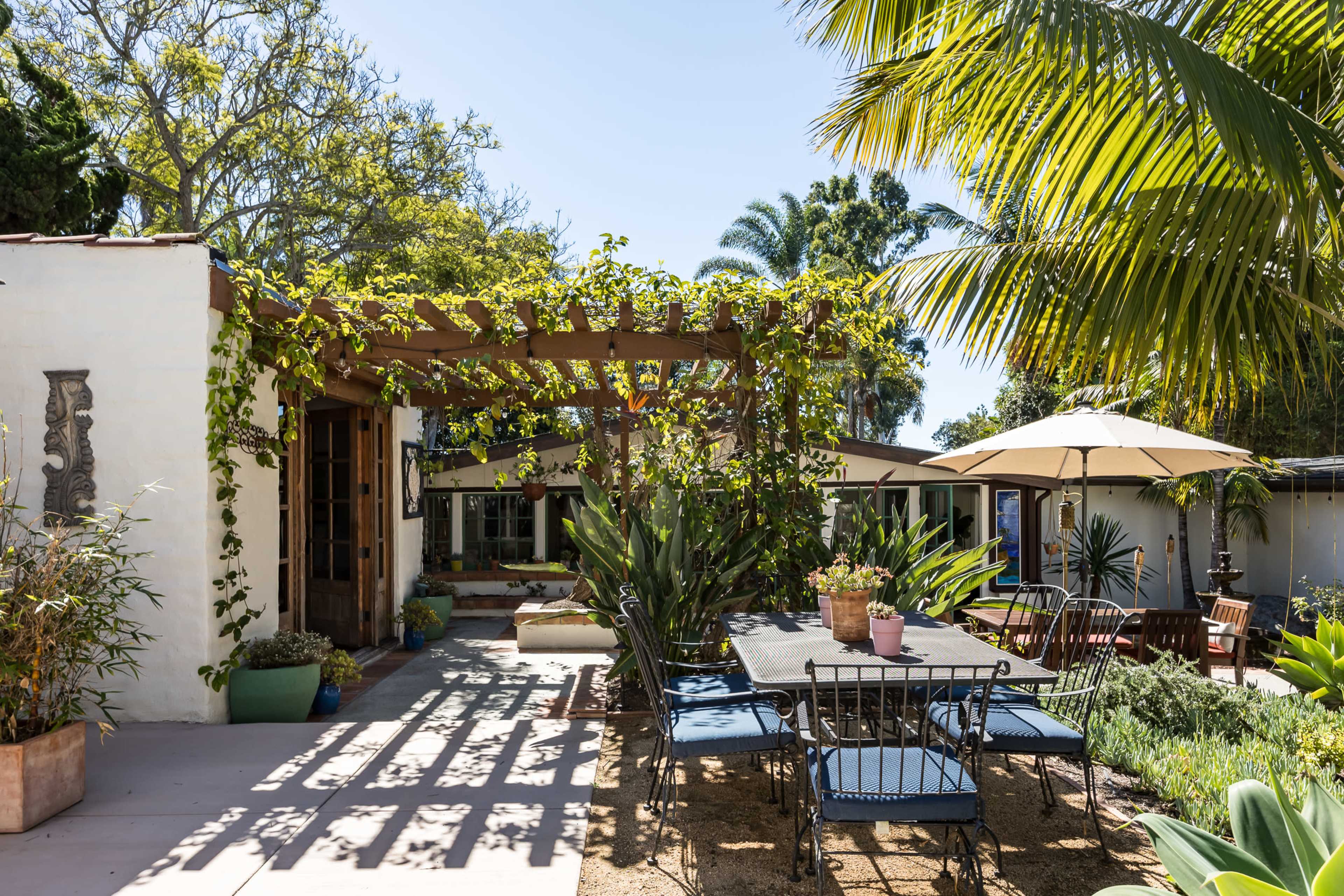 A sunlit outdoor patio features a large table surrounded by chairs, shaded by a pergola adorned with climbing vines, alongside landscaped greenery and palm trees.