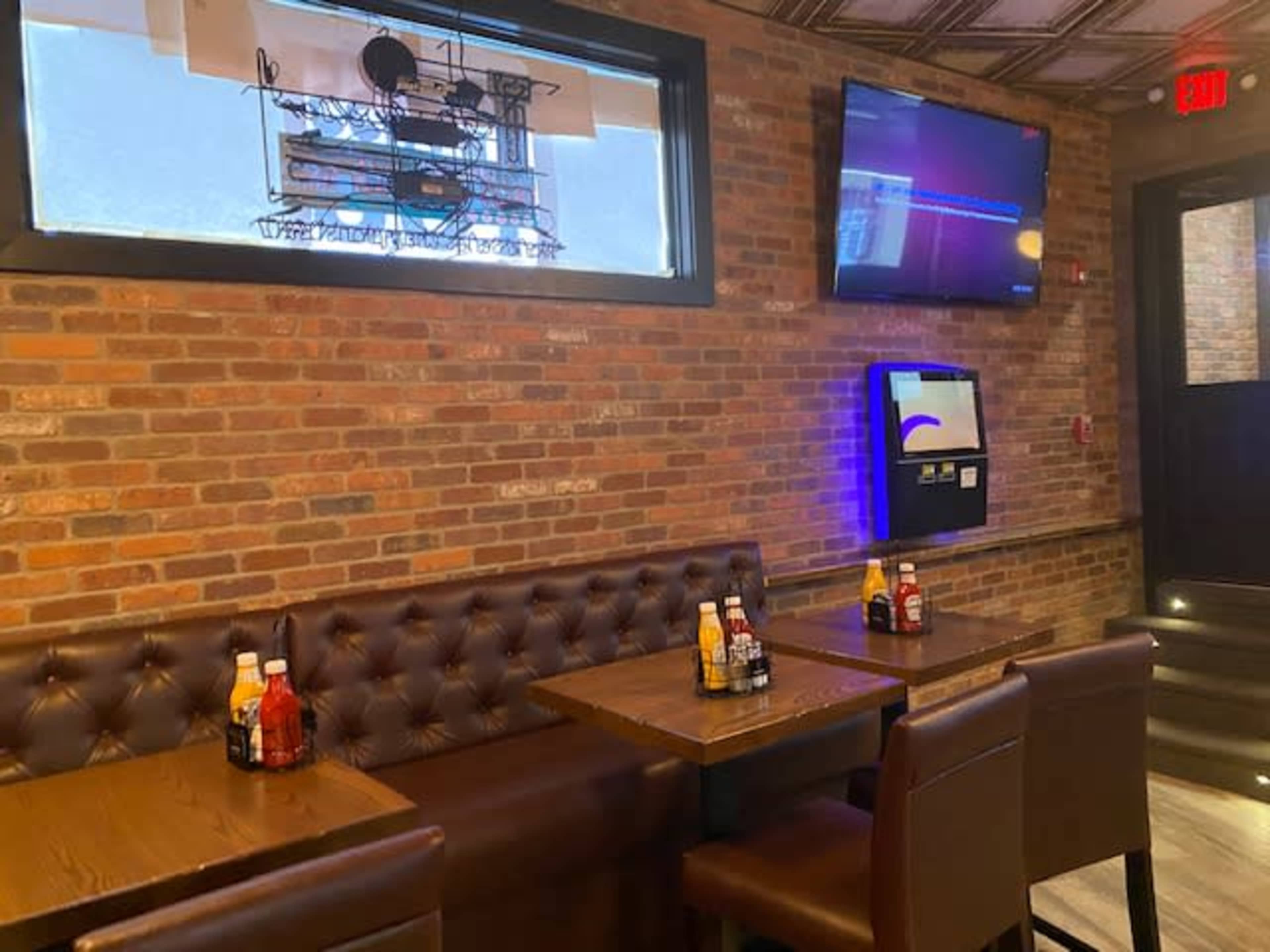 The image shows a seating area in a restaurant with brown leather booths, wooden tables, and condiments arranged on the tables against a brick wall.