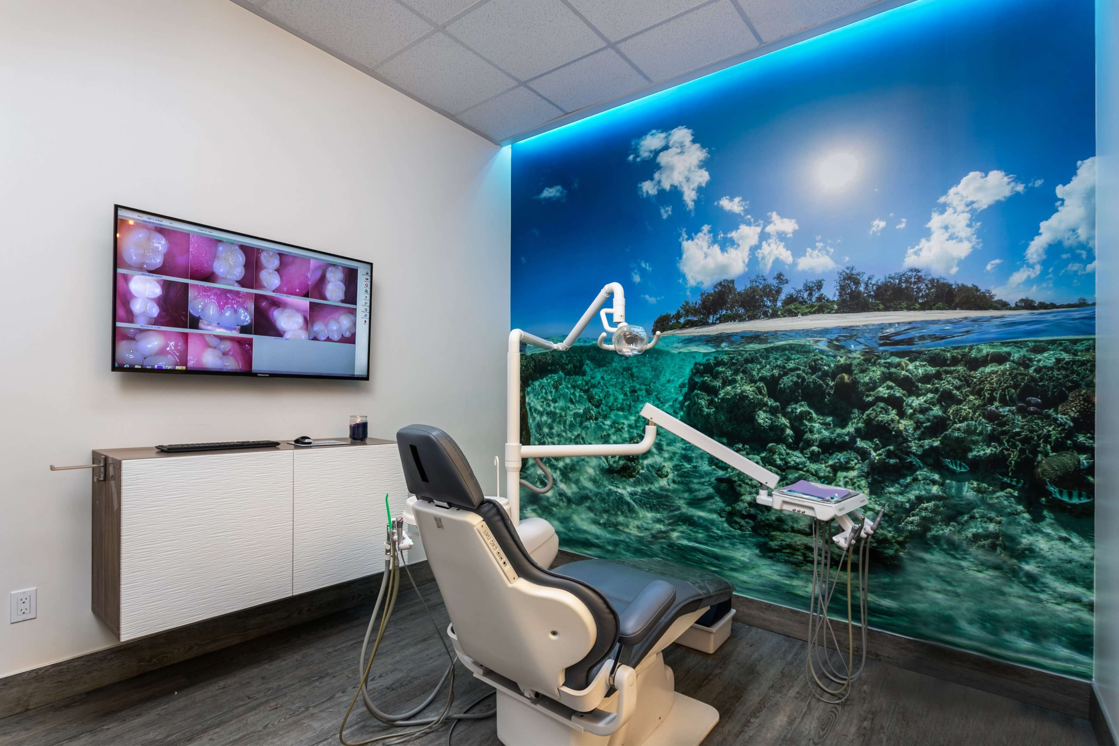 A dental examination room featuring a dental chair, a wall-mounted screen displaying dental images, and a large mural of a serene ocean landscape.