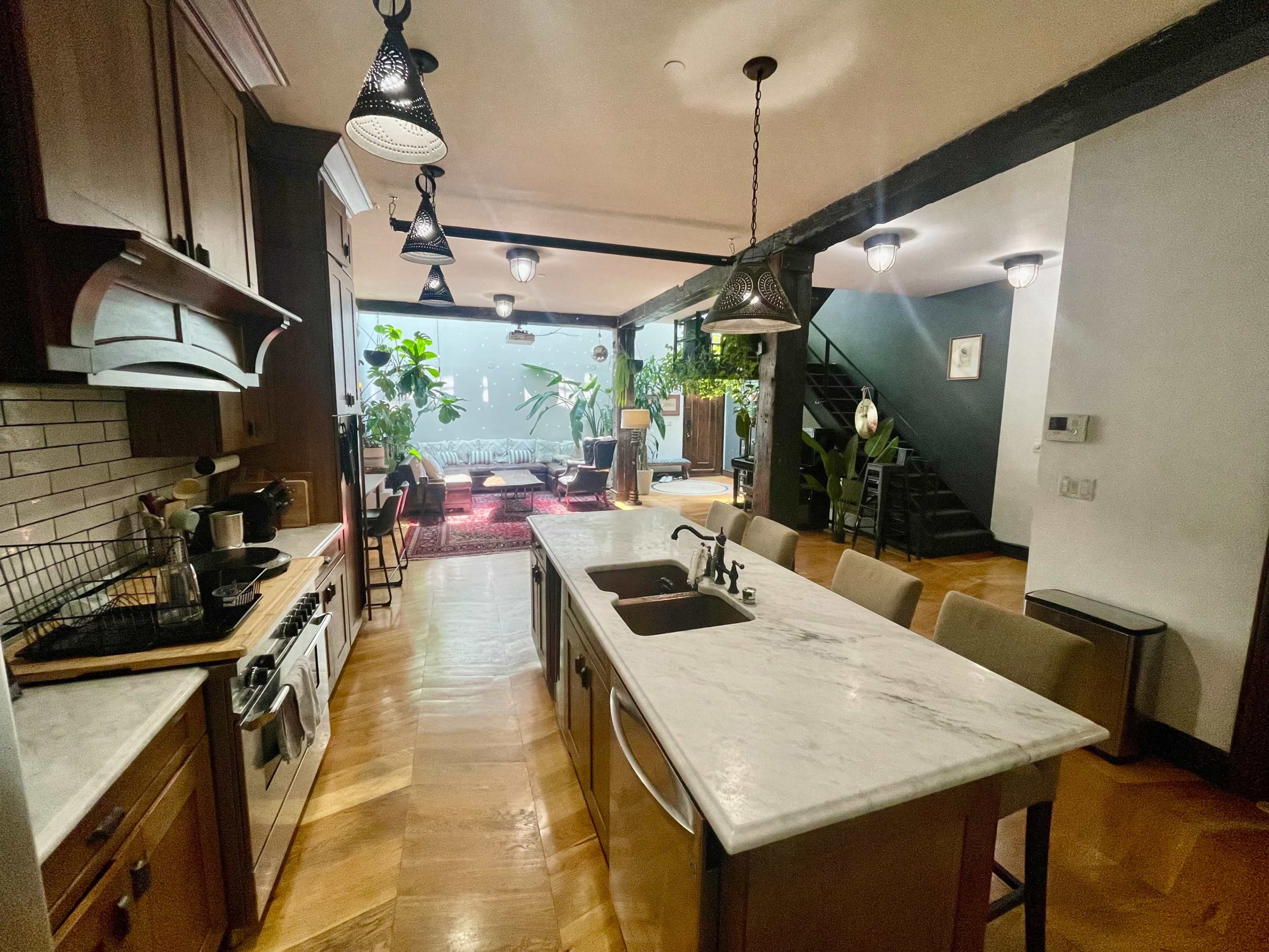A modern kitchen with a marble island, wooden cabinetry, and an open layout that leads to a plant-filled living area.