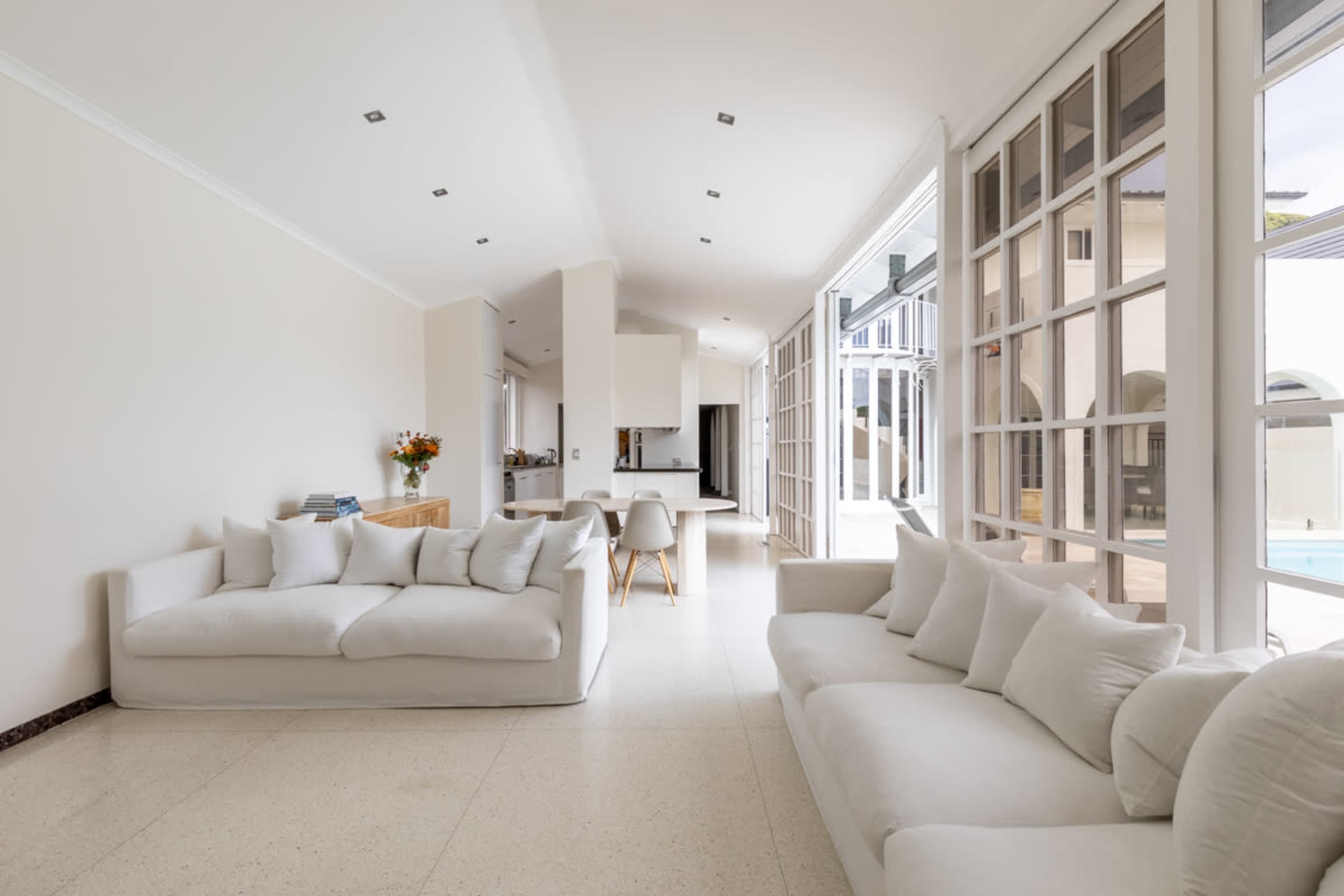 A spacious living room with two white sofas, large windows allowing natural light, and an open view of a kitchen area.