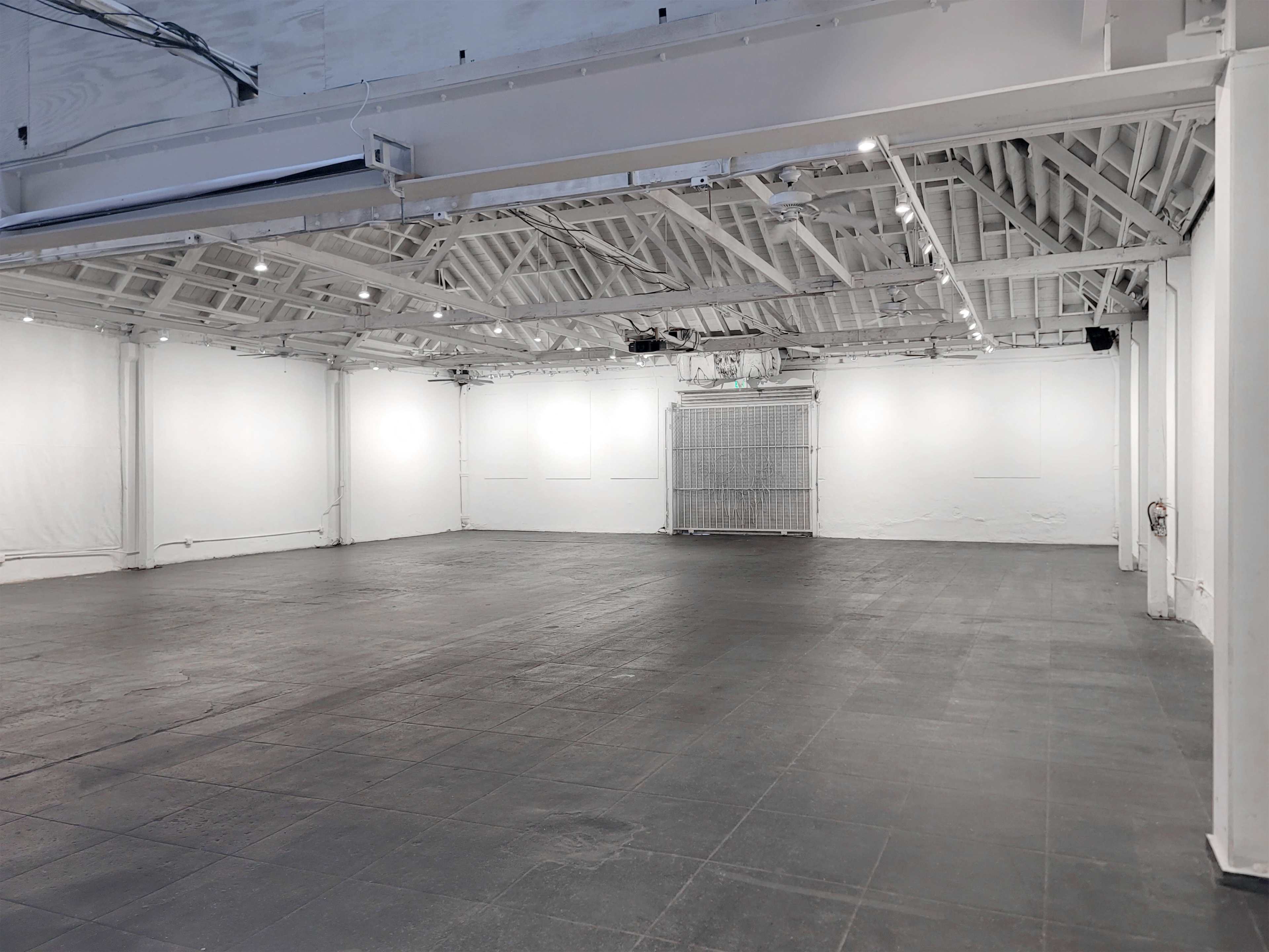 A large, empty white-walled interior space with a concrete floor and exposed wooden beams in the ceiling.