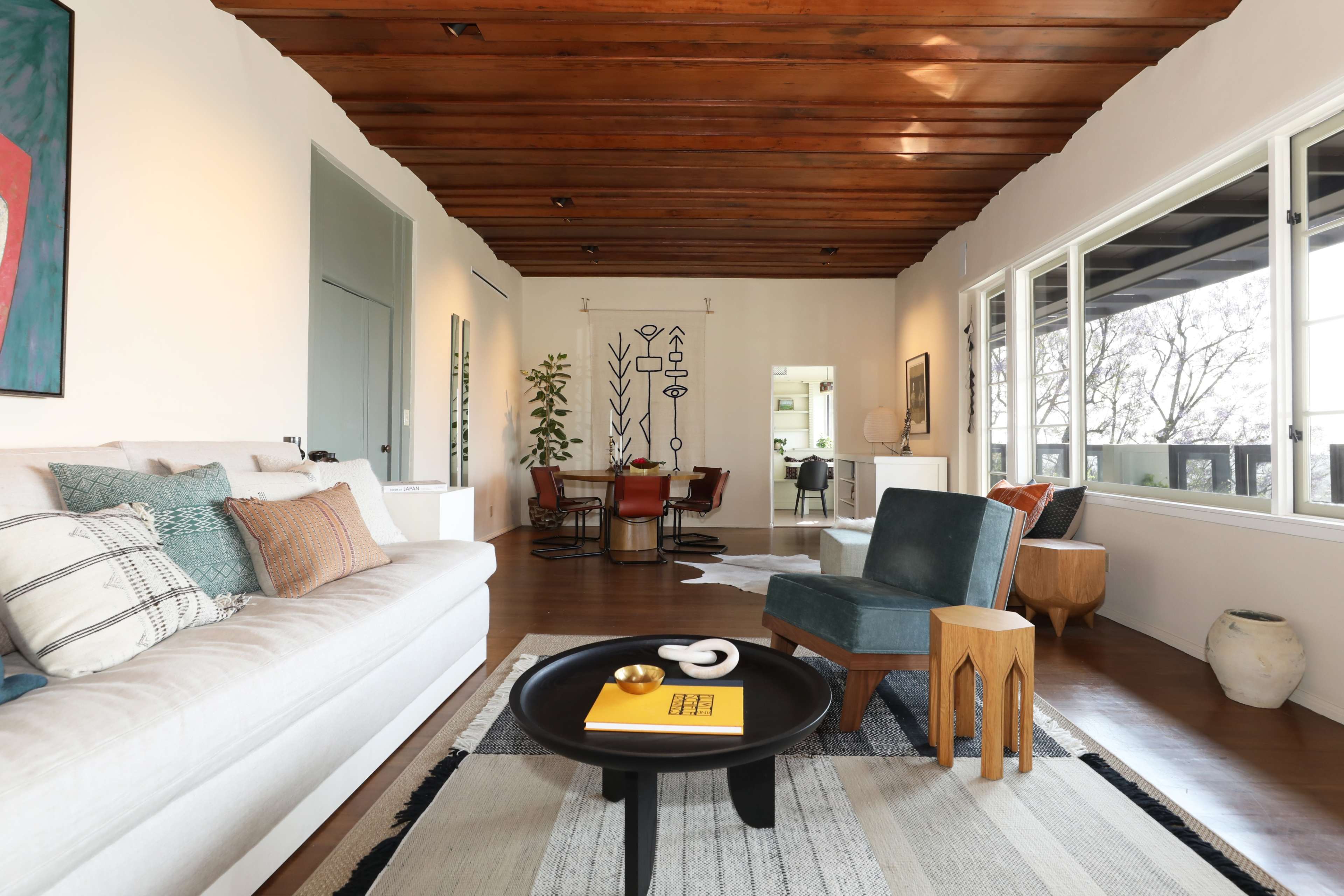 A modern living room with a light-colored sofa, wooden beam ceiling, and a coffee table, leading to an adjacent dining area with a large window.