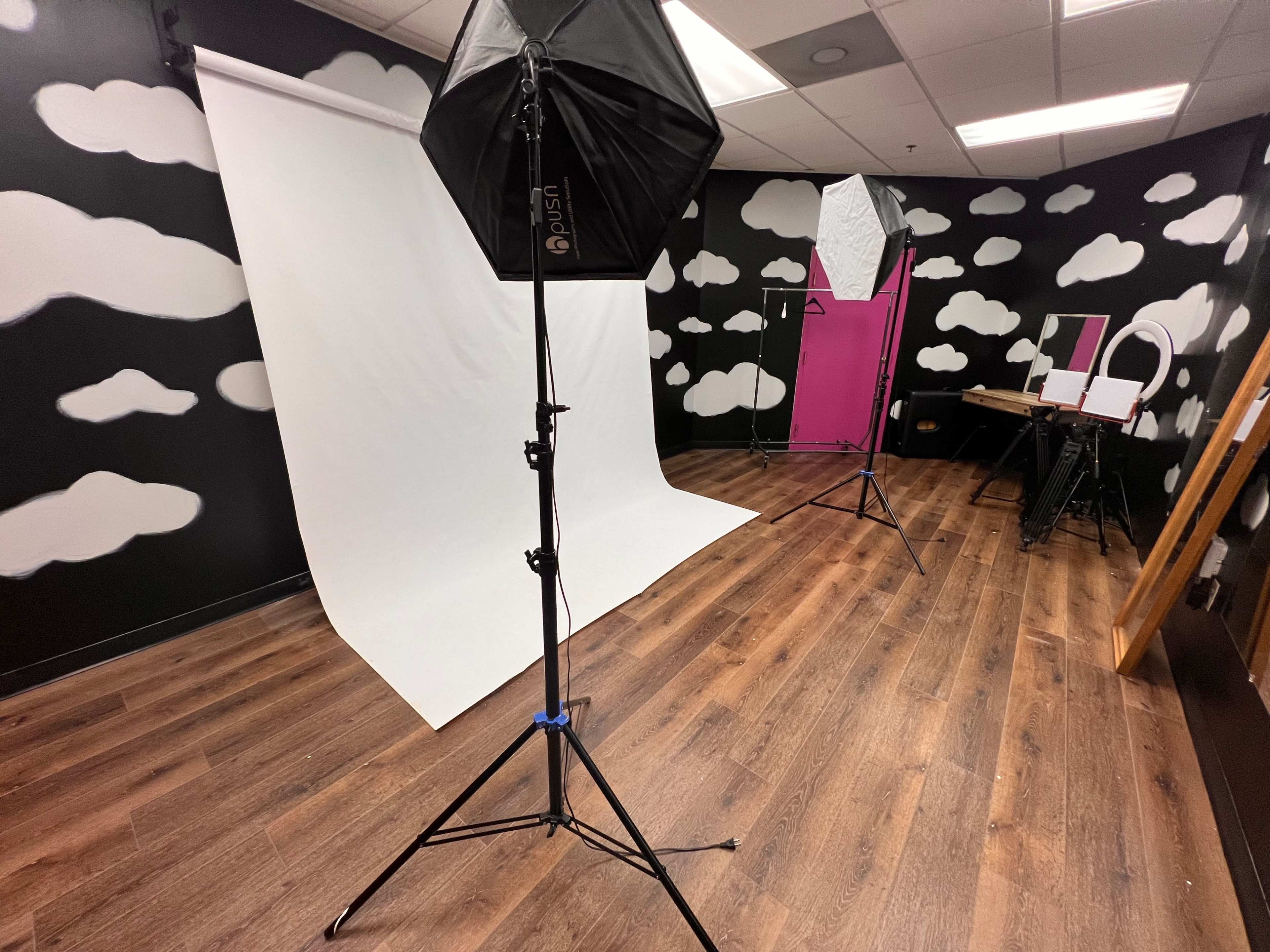 The image shows a photography studio featuring a white backdrop, softbox lights, and a black and white cloud-patterned wall.
