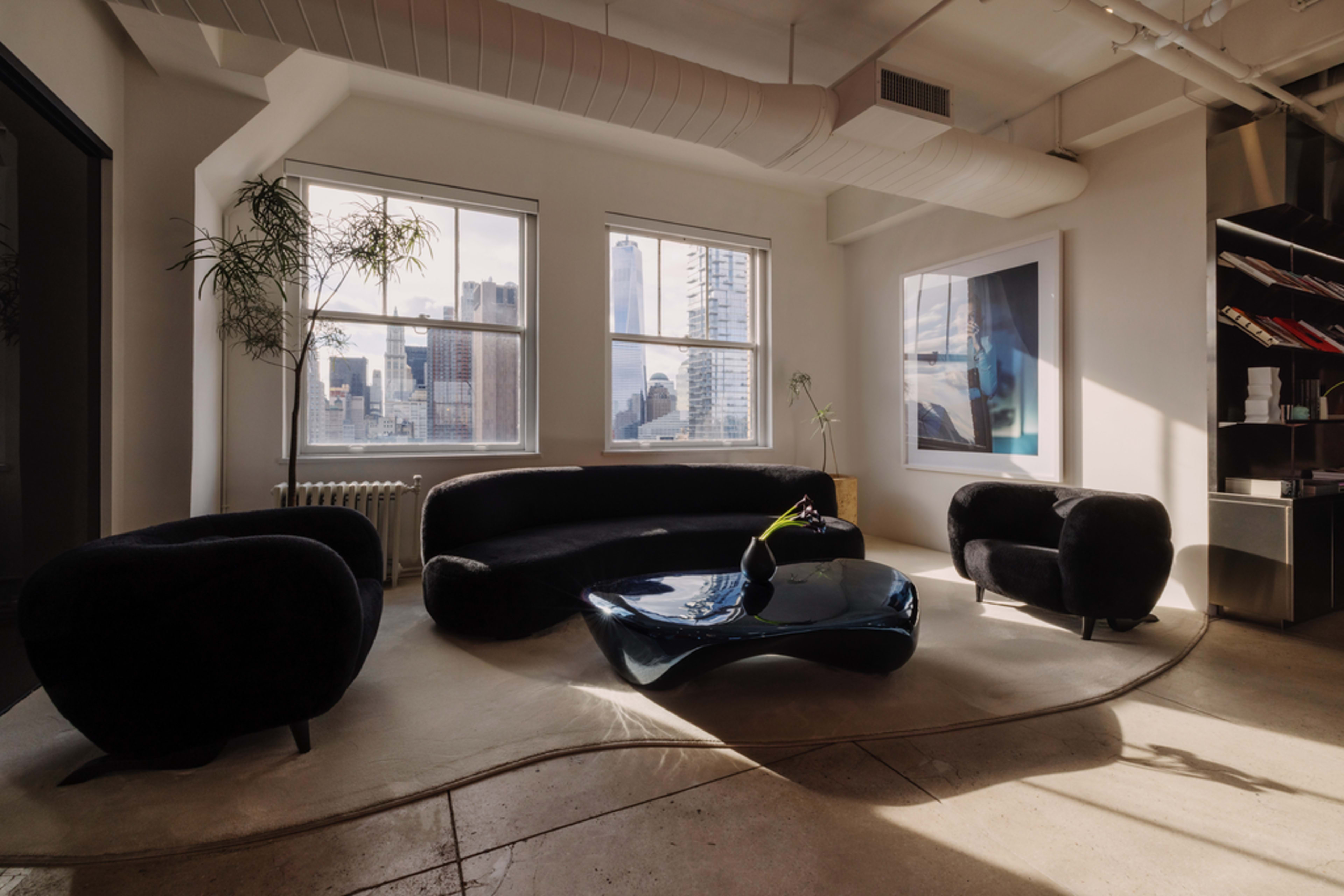 The image shows a modern living space featuring a curved black sofa, two round black chairs, and a unique blue coffee table, with large windows overlooking a city skyline.