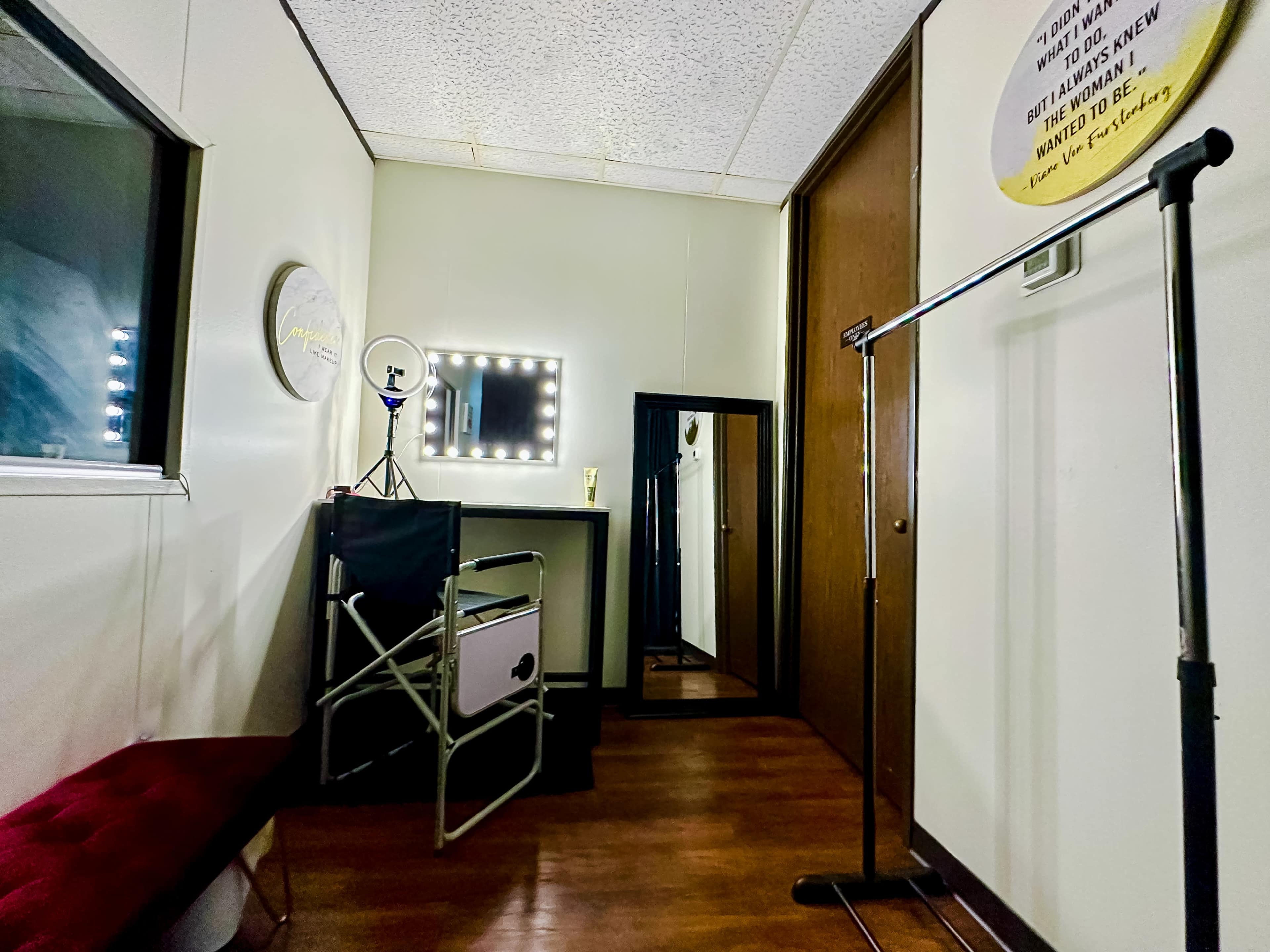A small, well-lit room with a mirror, a ring light, a folding chair, and a clothing rack against the walls.