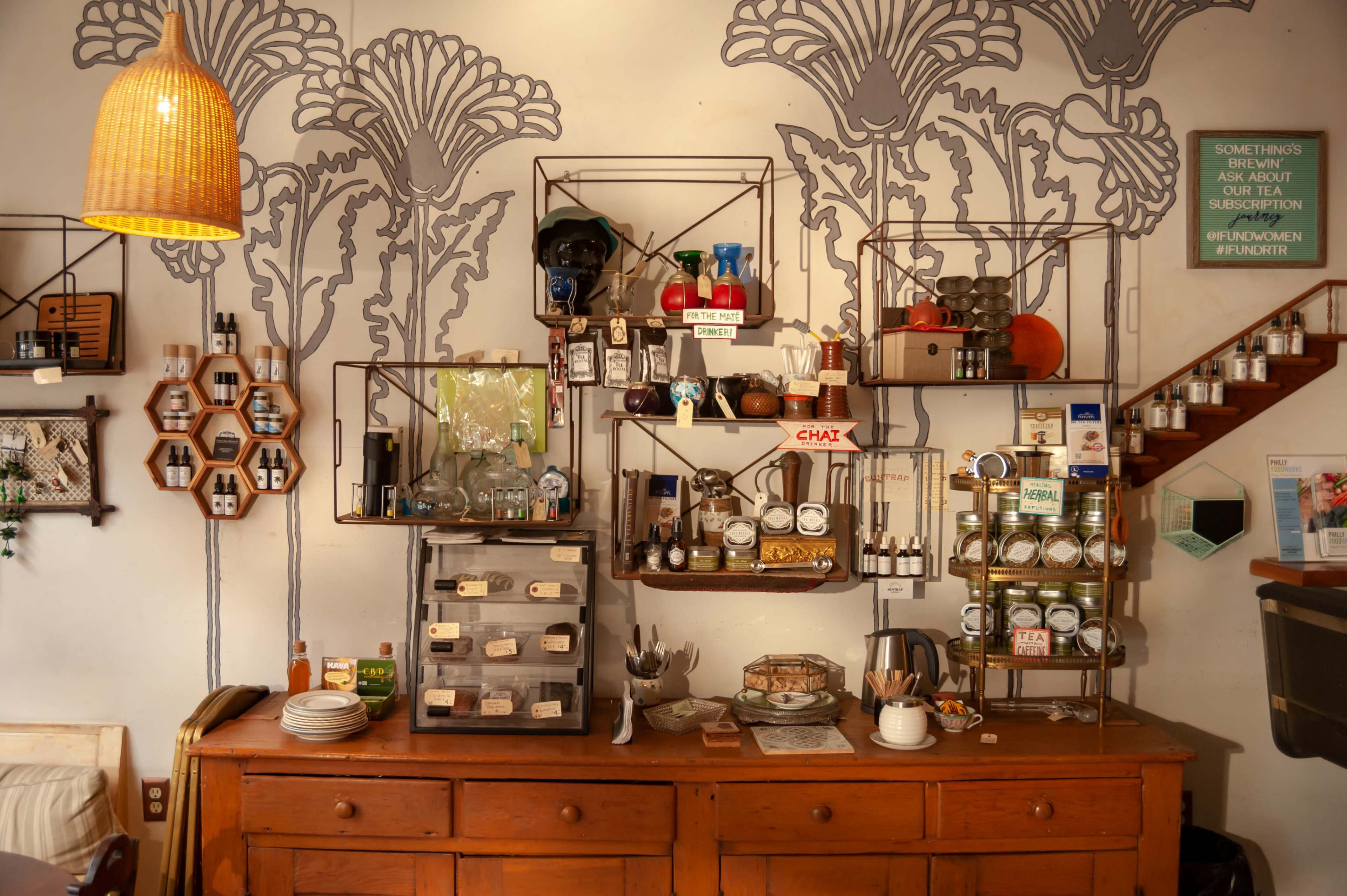 The image shows a cozy café interior with a wooden countertop displaying various food and drink items, shelves filled with jars and bottles, and decorative wall art featuring floral patterns.