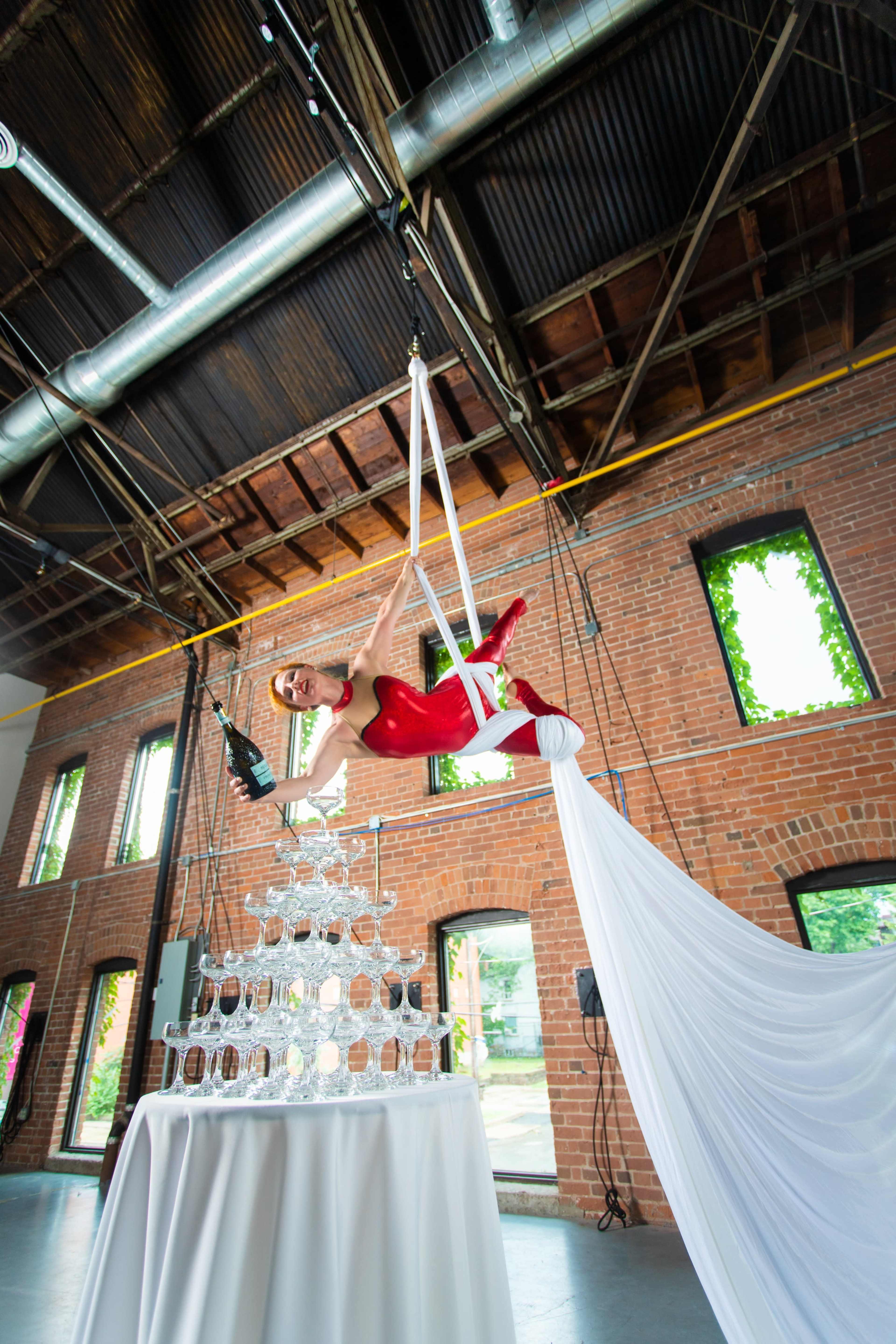 An aerial performer in a red outfit balances gracefully above a glass tower during a festive event in an industrial space.