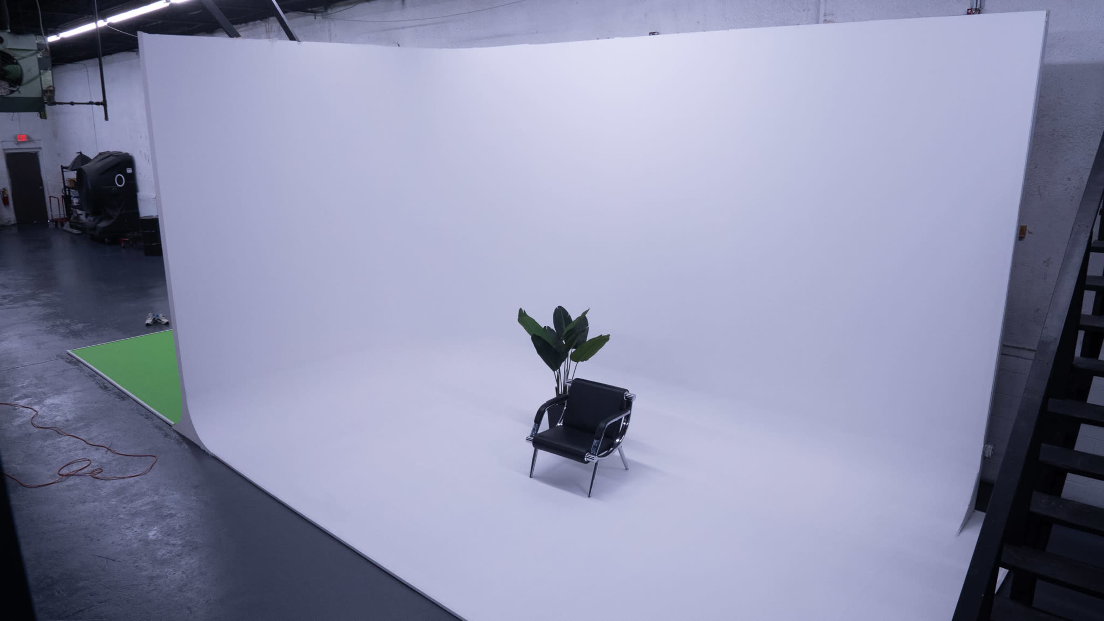 The image shows a minimalist studio setup with a single black chair and a potted plant placed on a white backdrop.