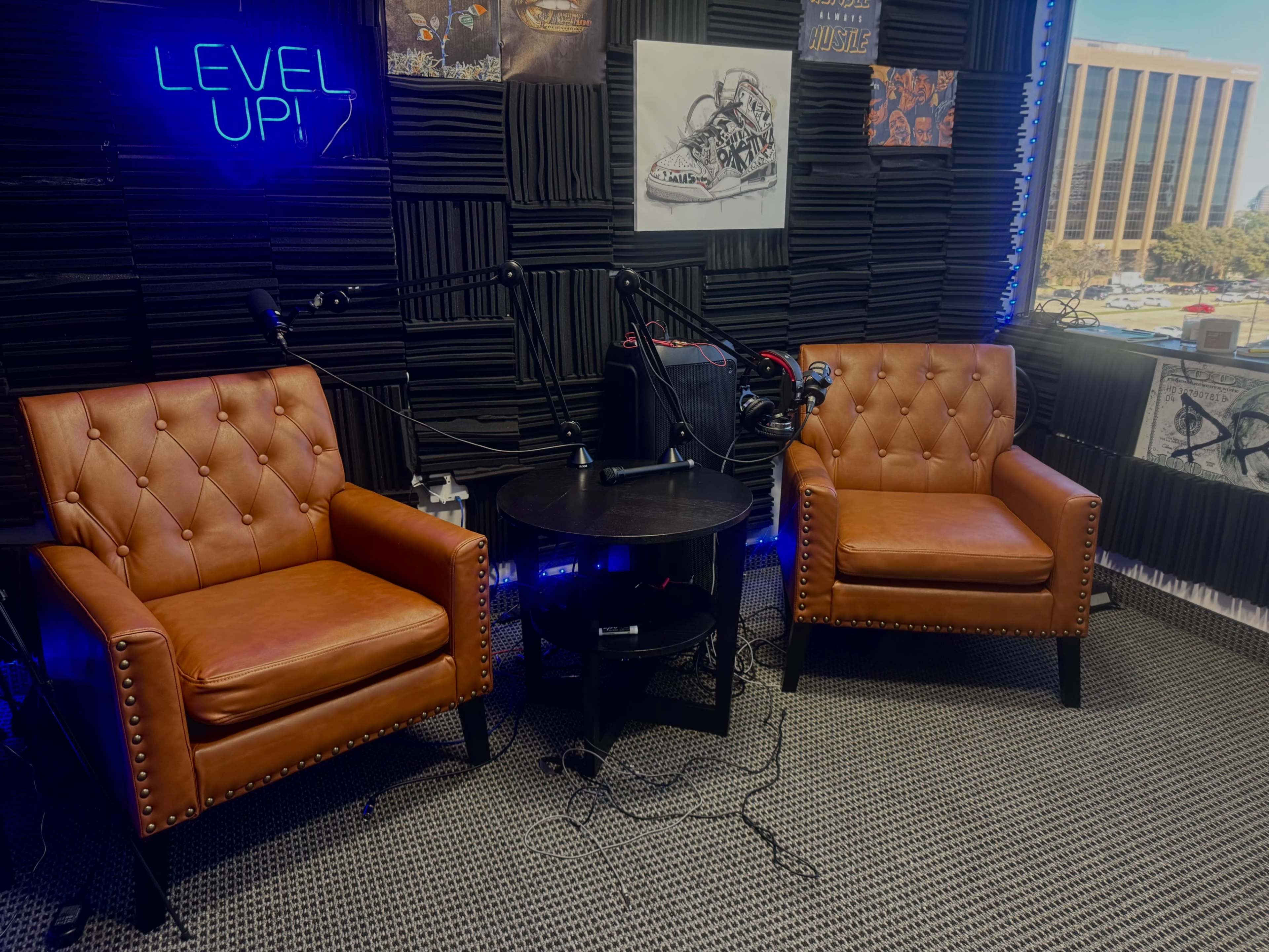The image shows a recording studio with two brown leather chairs, a small round table, and soundproof black panels on the walls.