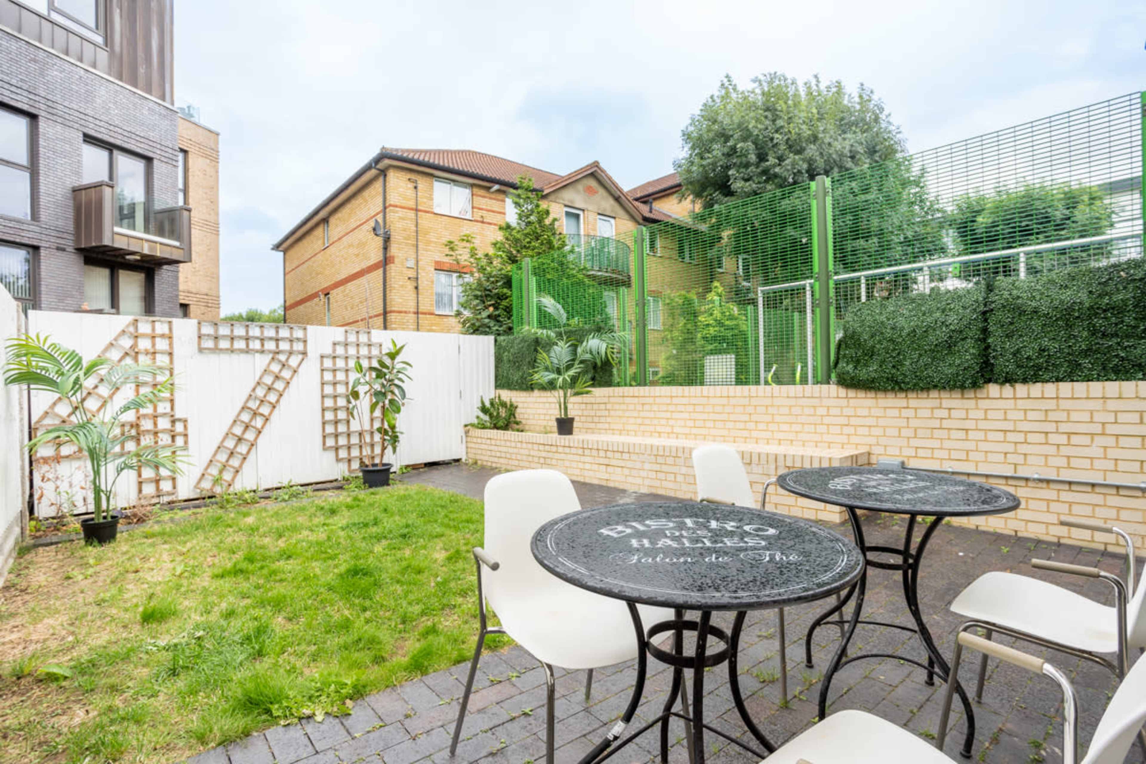 A small outdoor courtyard with two round tables and chairs, bordered by green hedges and residential buildings in the background.