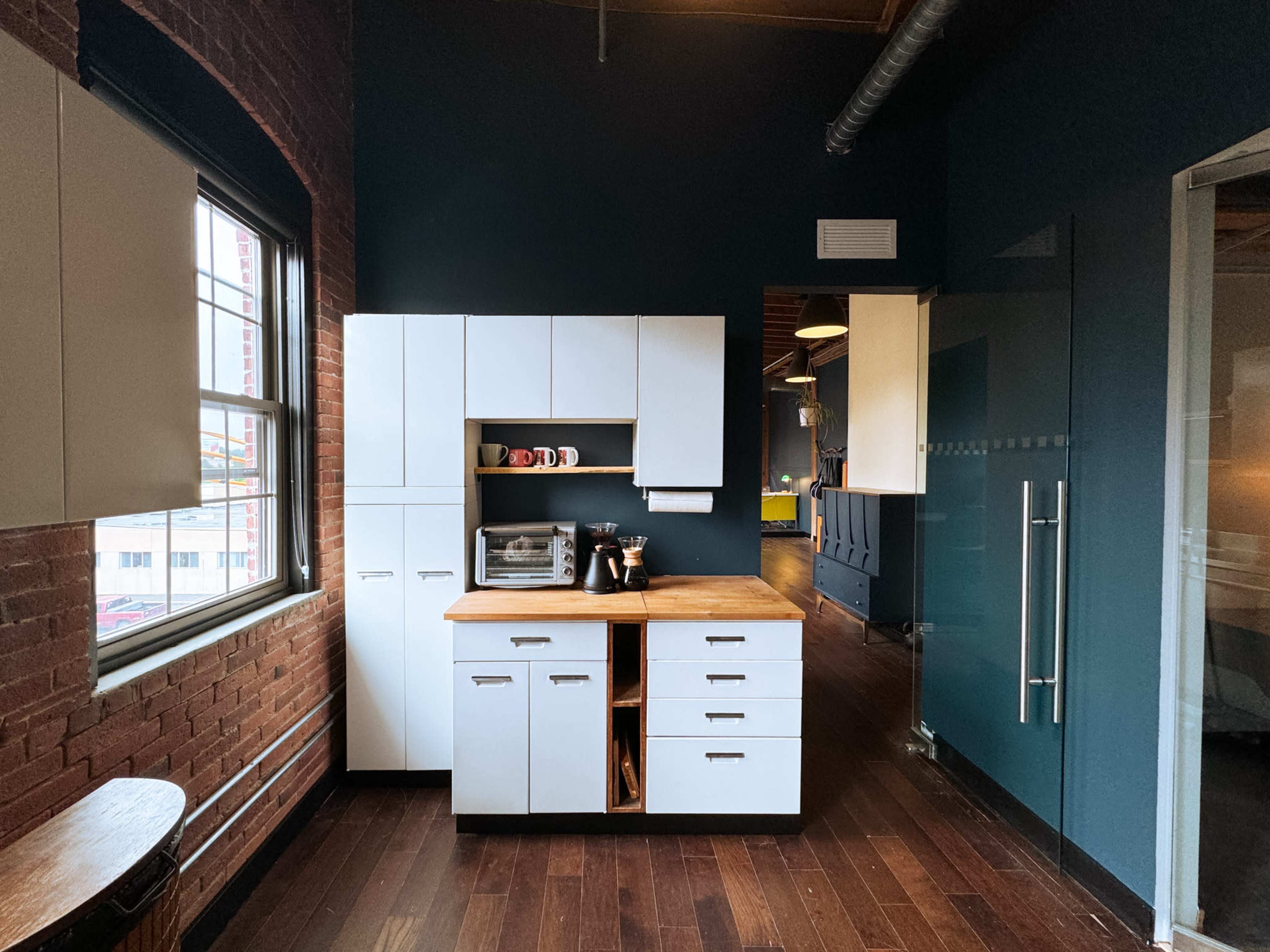 The image shows a modern kitchen with dark blue walls, wooden flooring, and a white cabinetry unit that includes a countertop and small appliances.