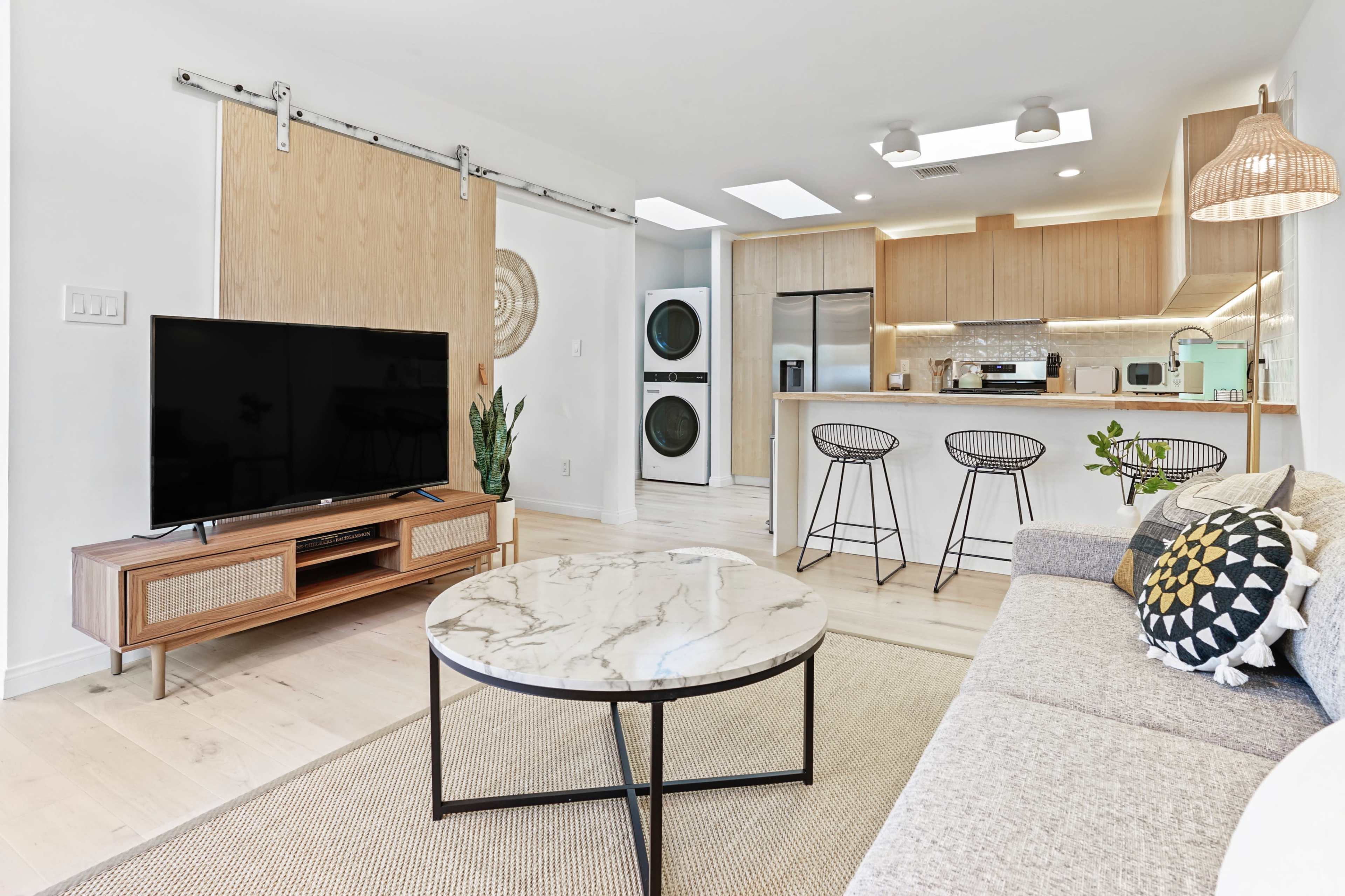 A modern living room features a television on a wooden console, a round marble coffee table, and a sectional sofa, with a kitchen area visible in the background.
