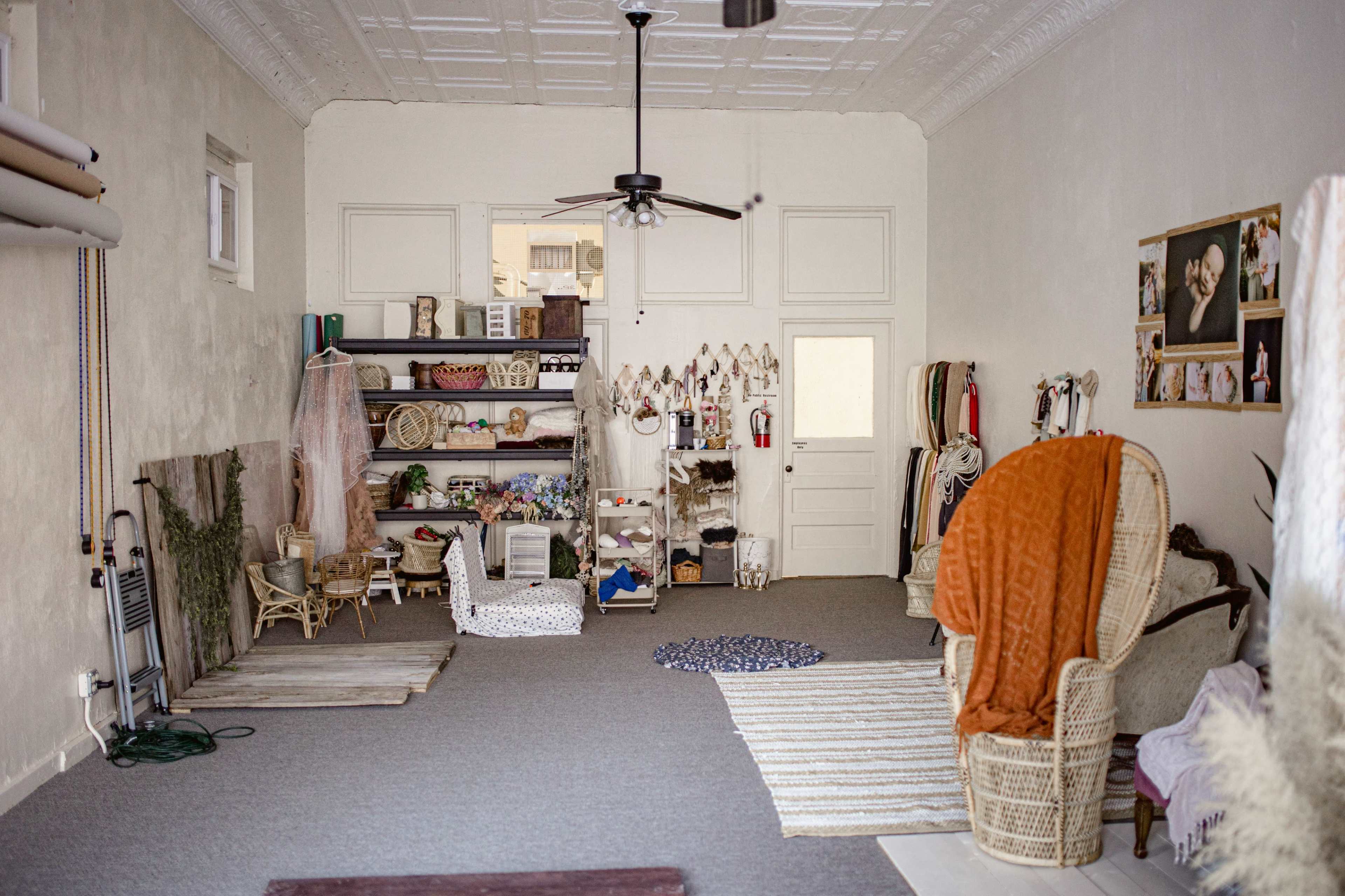 The image shows a spacious, well-lit room with a mix of furniture including a chair, shelves filled with decorative items, and several textiles on the floor.