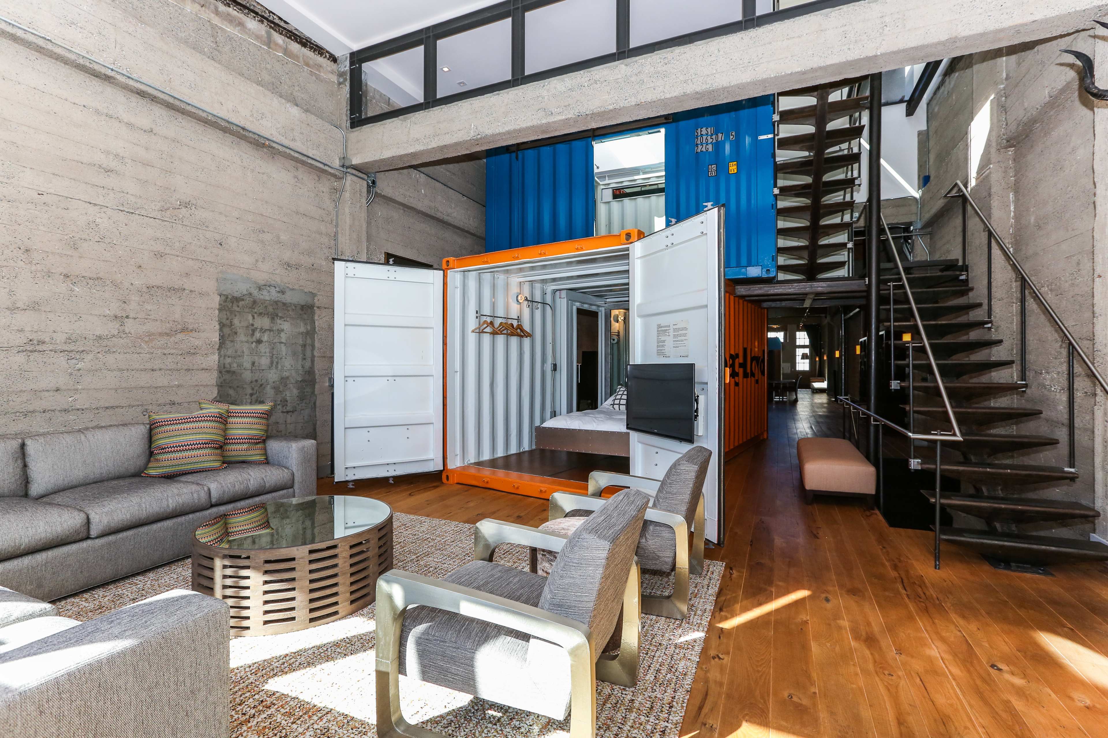 A modern interior space featuring a living area with a seating arrangement, a glass coffee table, and a shipping container used as a bedroom, with a staircase leading to an upper level.
