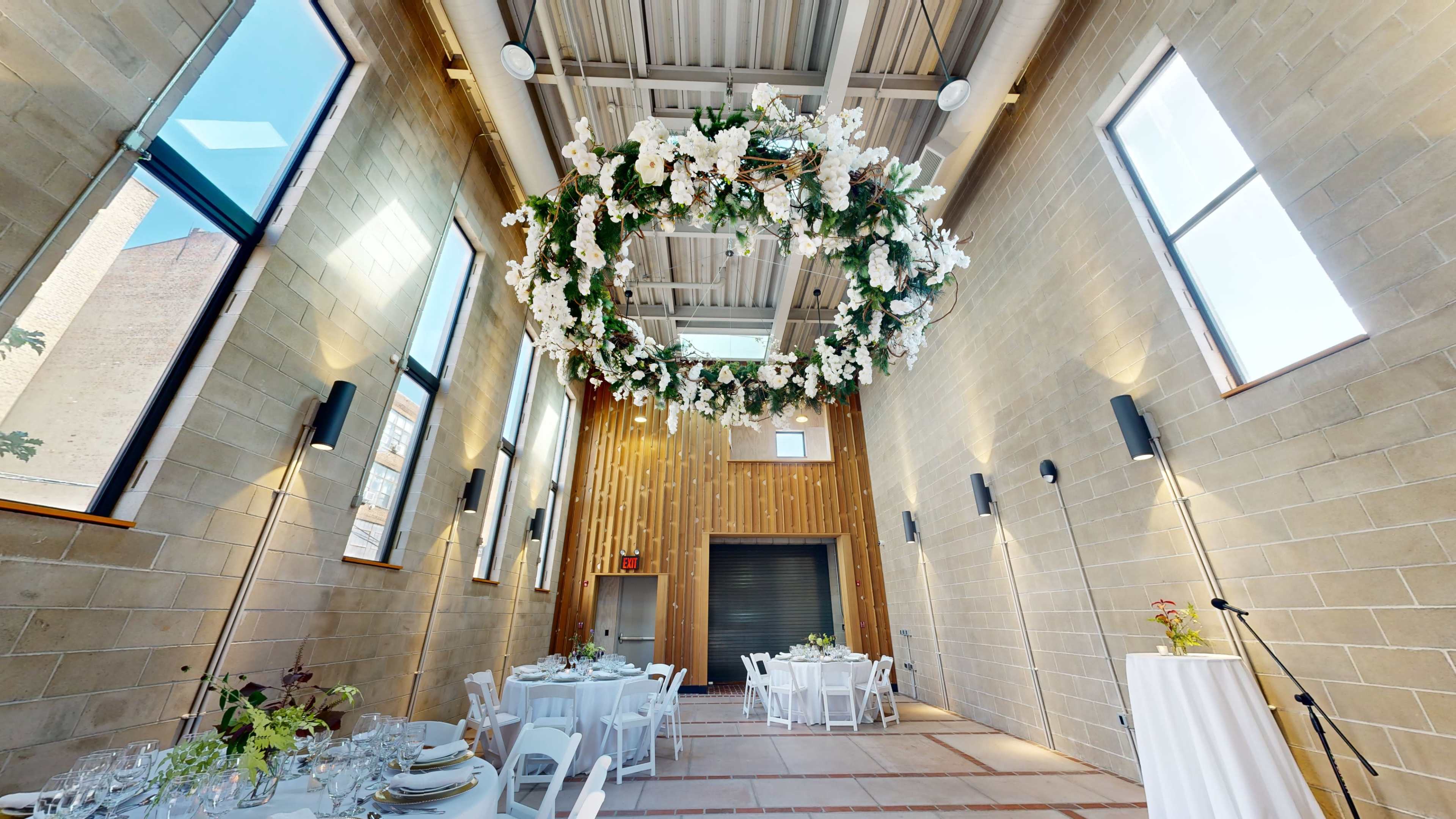 The interior of a modern venue features tables set for dining beneath a large floral chandelier, with windows allowing natural light to fill the space.