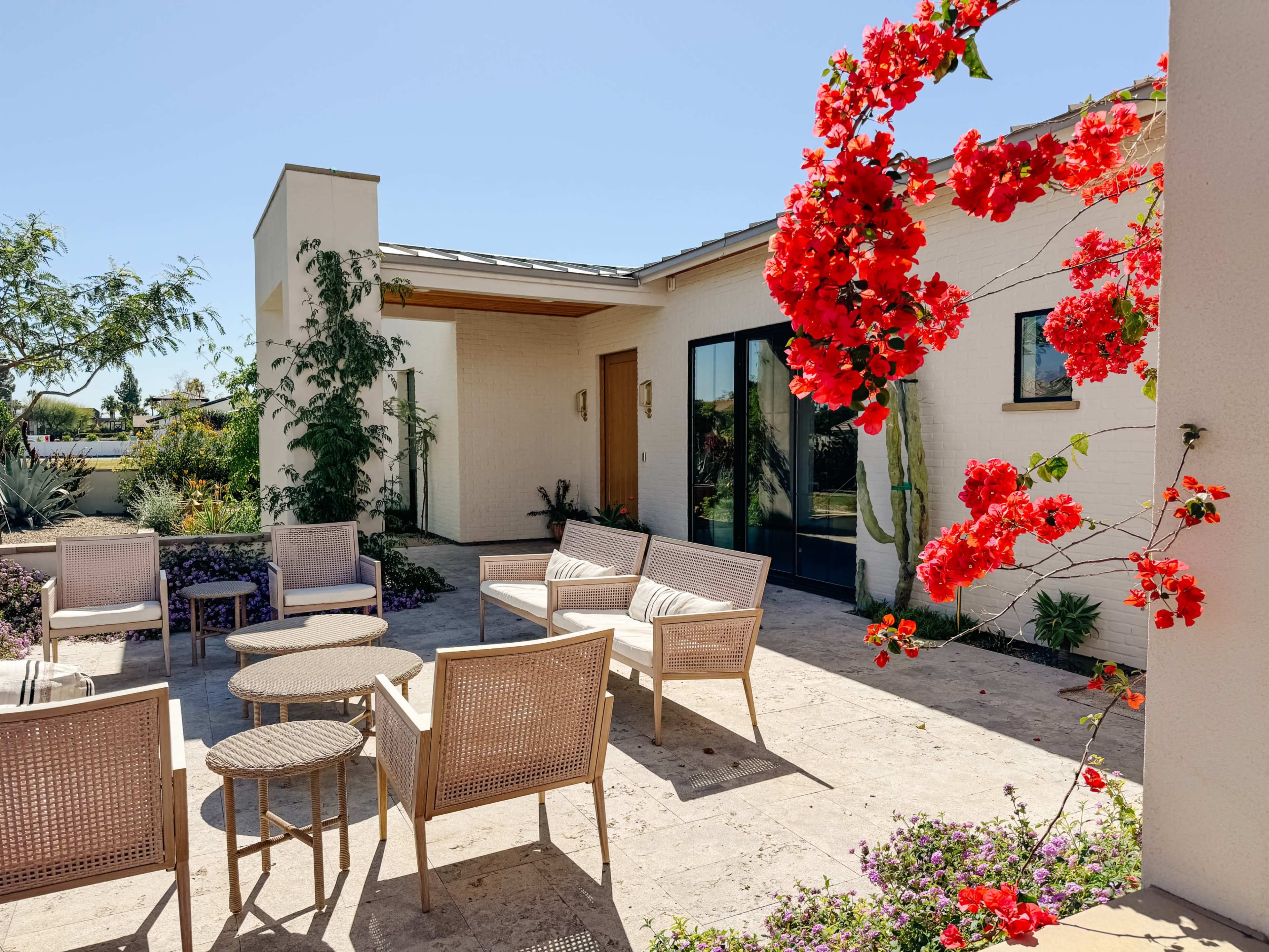 A patio area features rattan furniture arranged around circular tables, framed by vibrant red bougainvillea and a modern white house in the background.