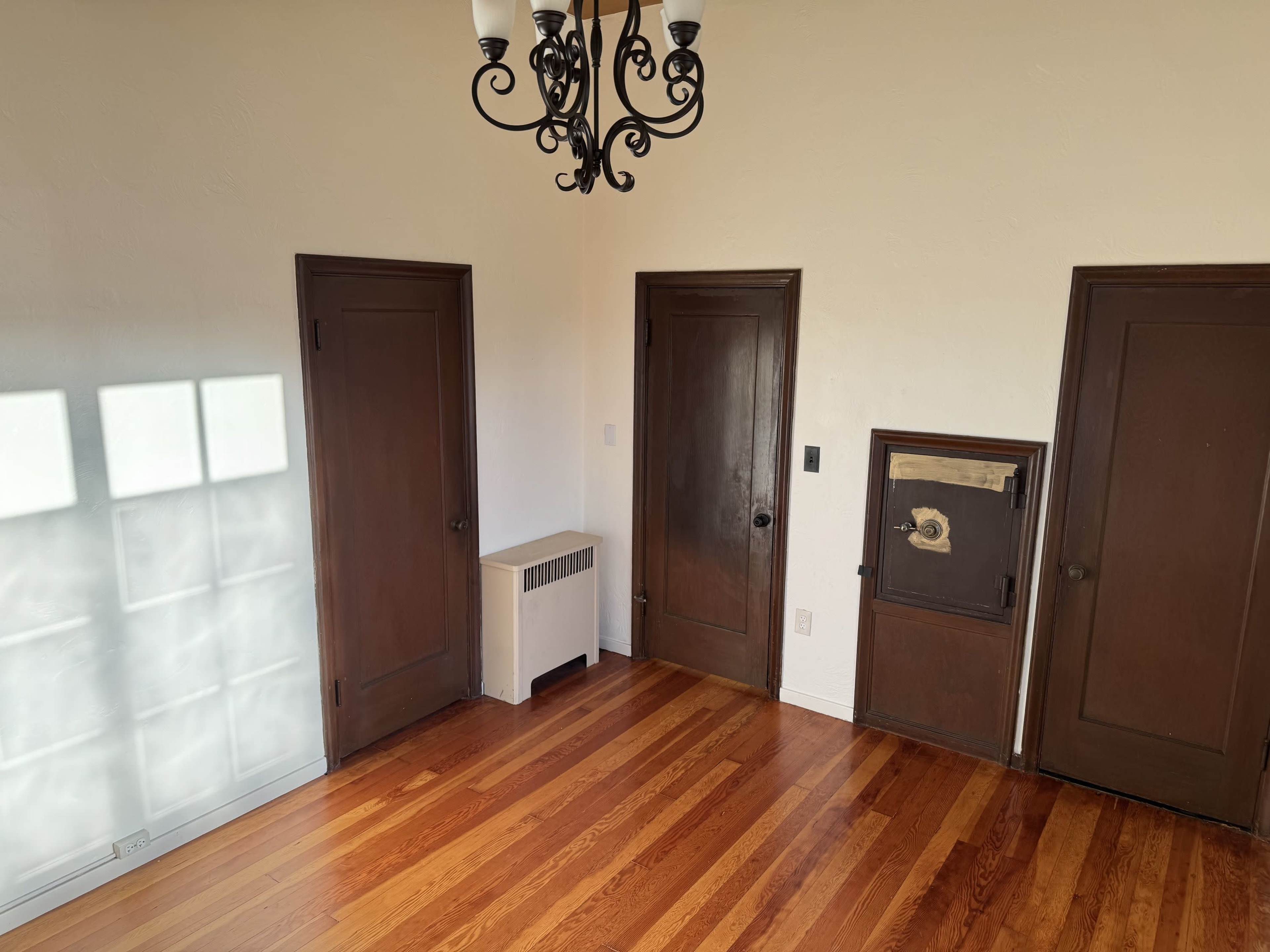 The image shows a room with hardwood floors, three brown doors, a wall heater, and a chandelier.