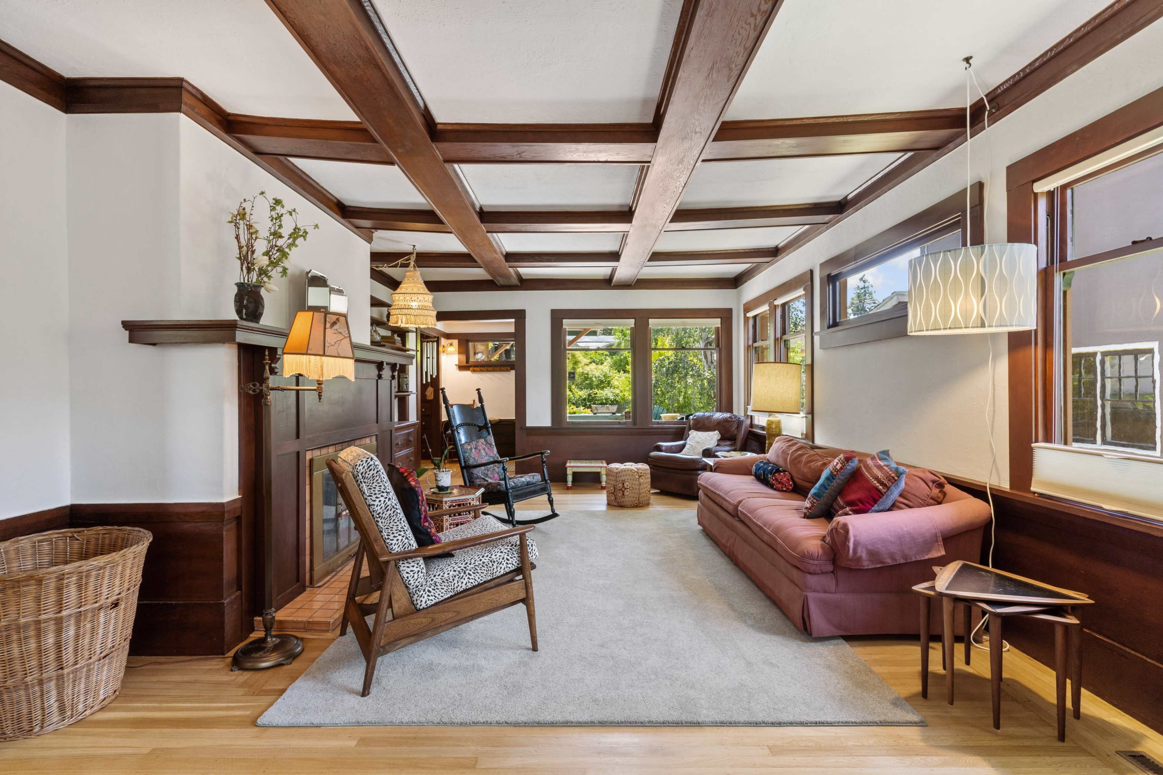 A cozy living room with wooden beams on the ceiling, a brown sofa, and a rocking chair, along with large windows allowing natural light to enter.