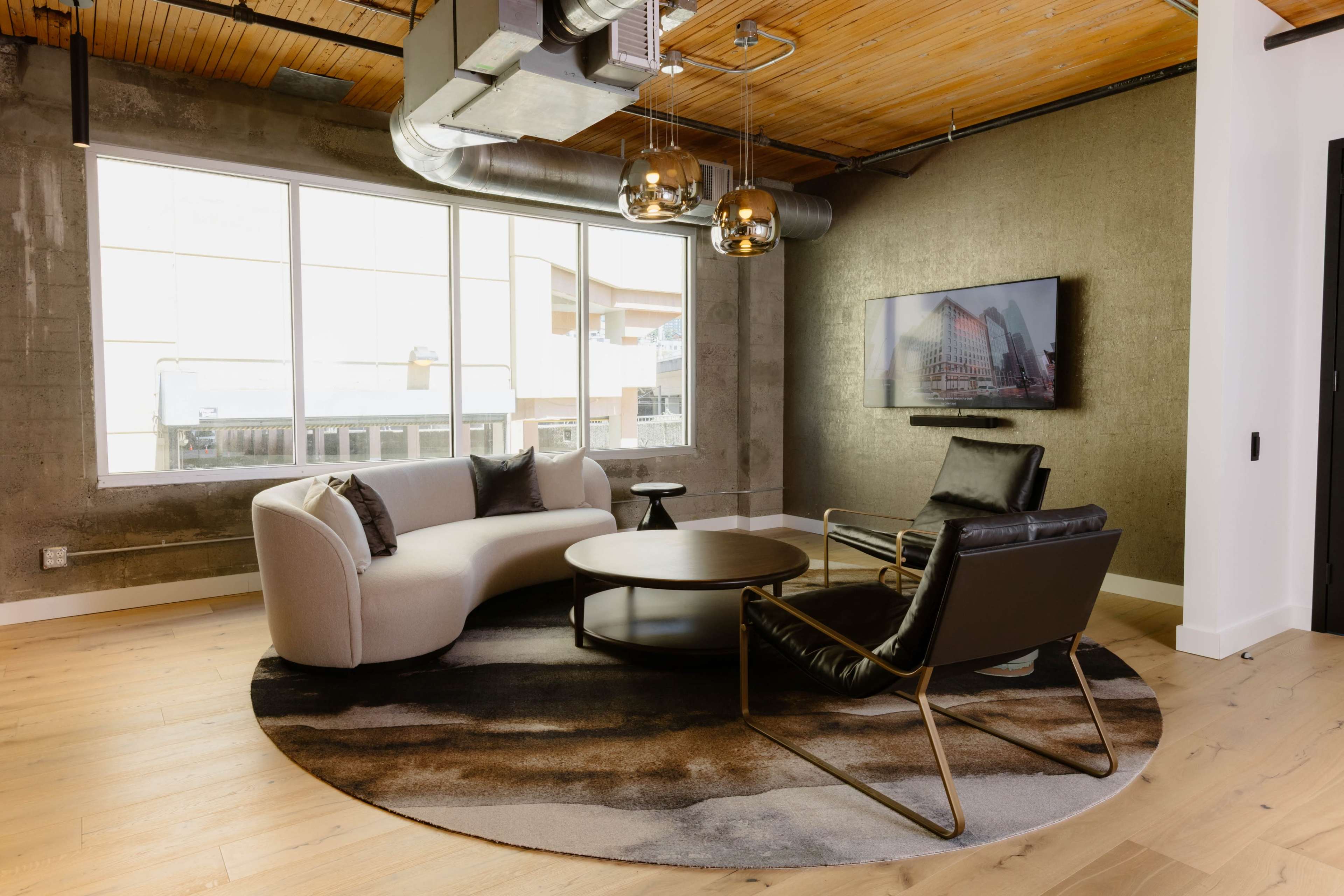The image shows a modern lounge area featuring a curved white sofa, a black leather chair, and a round coffee table on a circular rug, with large windows and a television mounted on the wall.